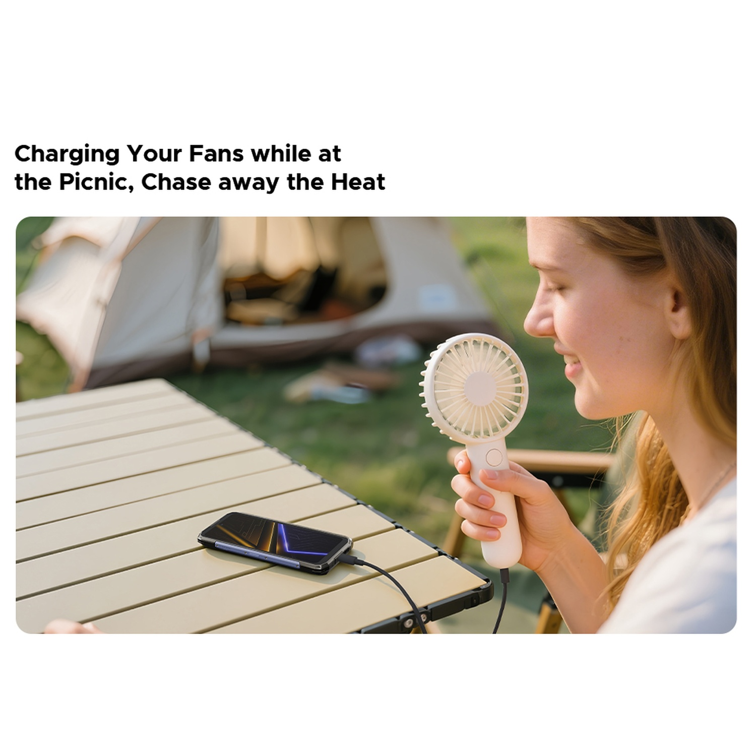 Una donna tiene in mano un ventilatore bianco e sorride. Un telefono si carica su un tavolo. Una tenda è sullo sfondo.