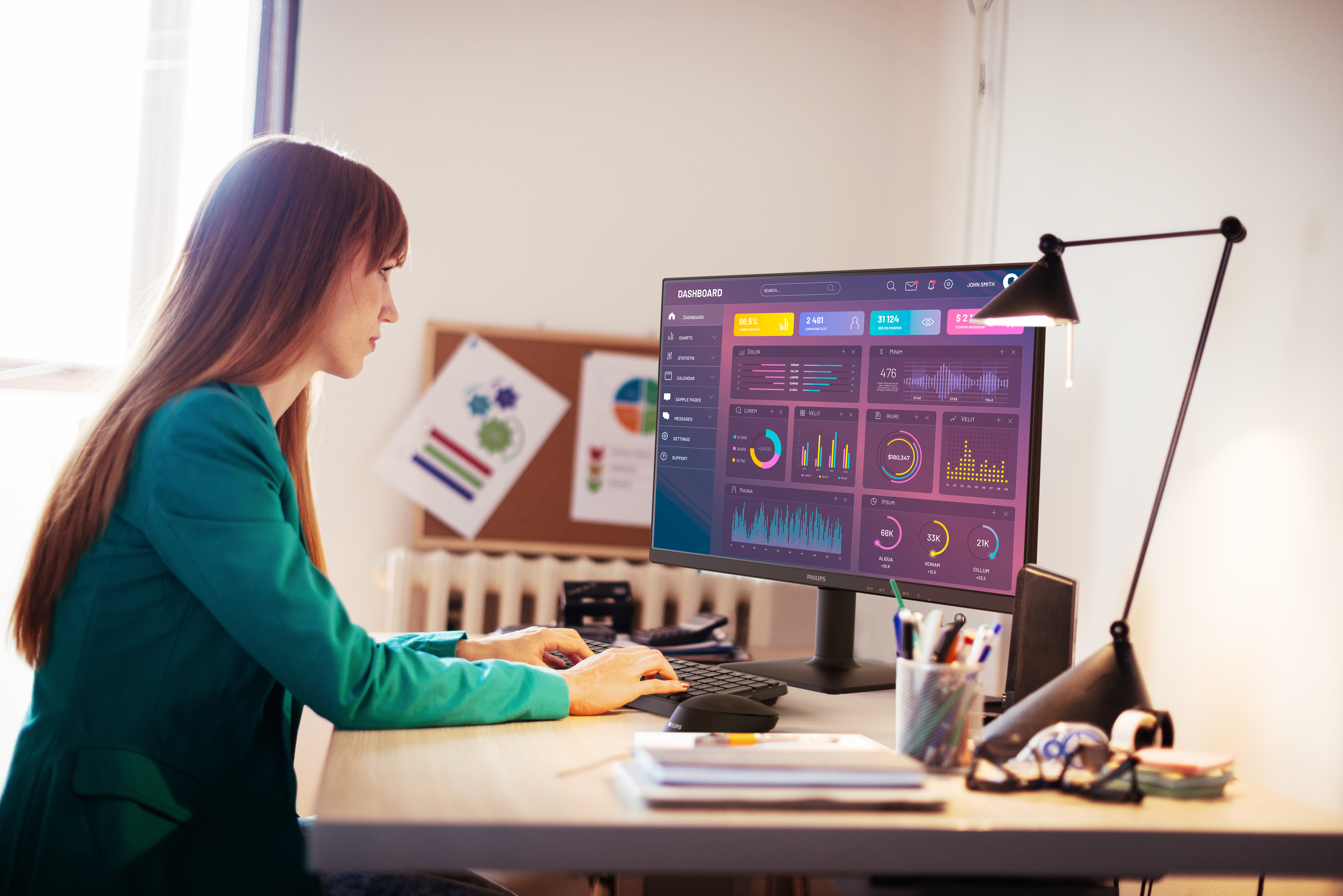 Eine Frau in einer teal Jacke arbeitet an einem Computer, der ein Dashboard anzeigt. Auf dem Schreibtisch befinden sich eine Lampe und Büromaterialien.