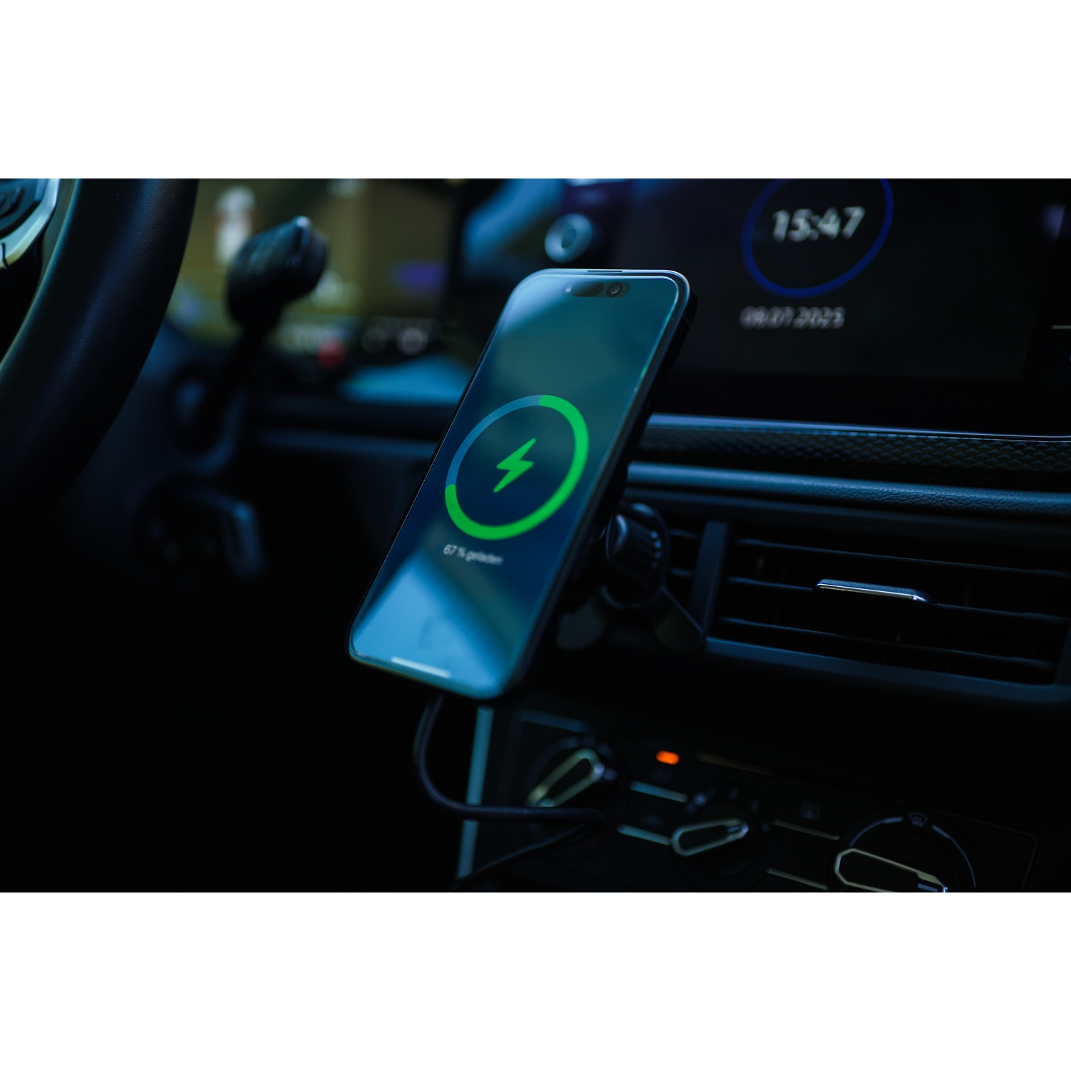 Uno smartphone in carica in auto mostra un'icona a forma di fulmine. Lo schermo mostra il 67% di carica. Sono visibili cruscotto e volante.