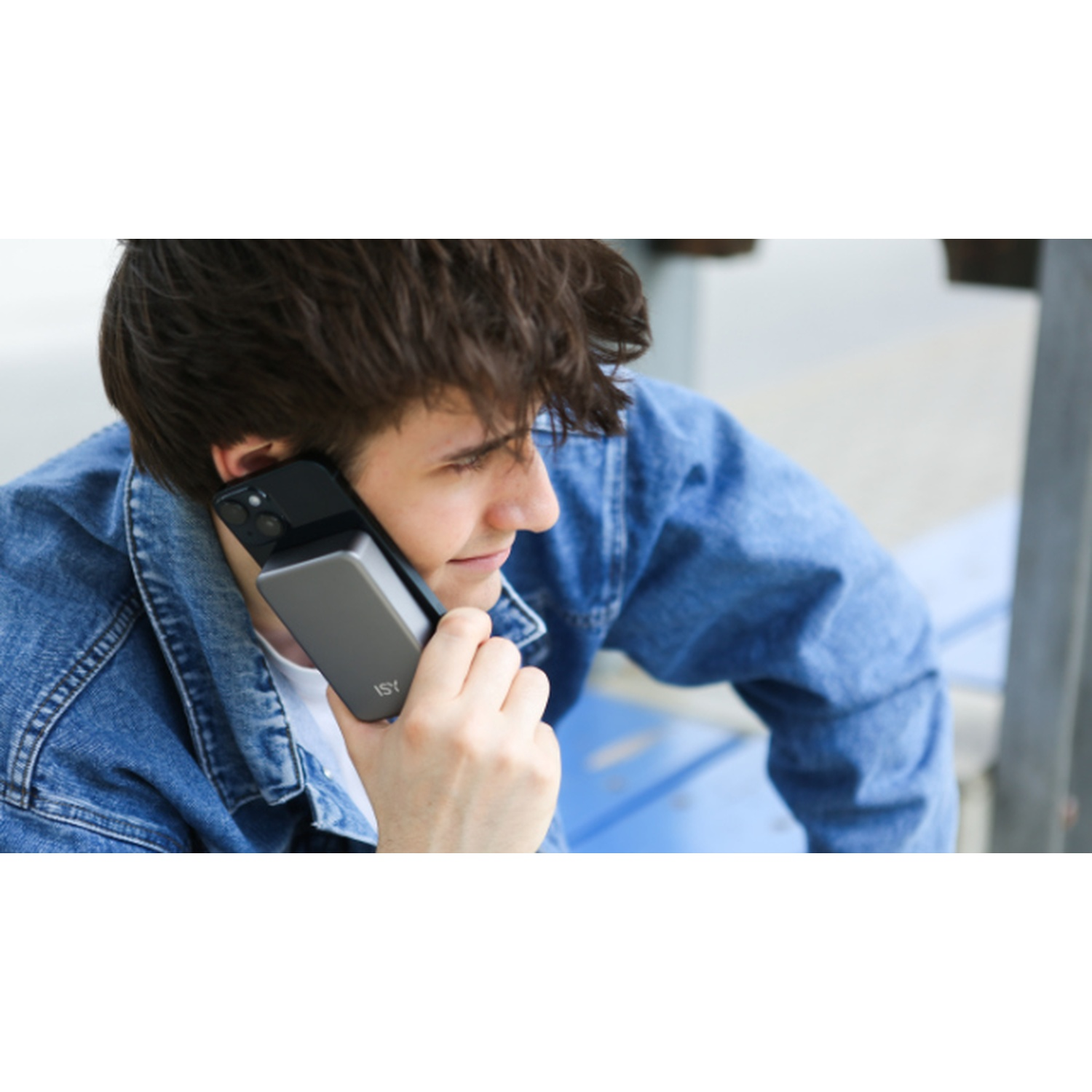 Un giovane con una giacca di jeans tiene un telefono e un power bank all'orecchio. Lo sfondo è sfocato.