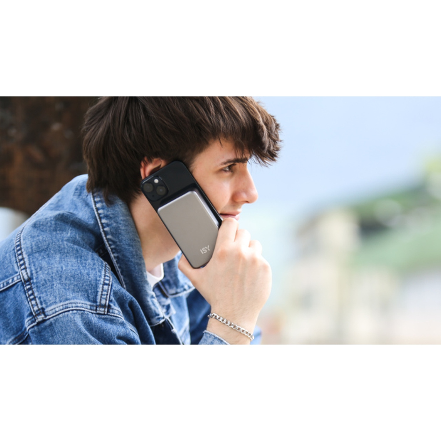 Un giovane con una giacca di jeans tiene un telefono all'orecchio. Il telefono è nero e argento. Indossa un braccialetto al polso.