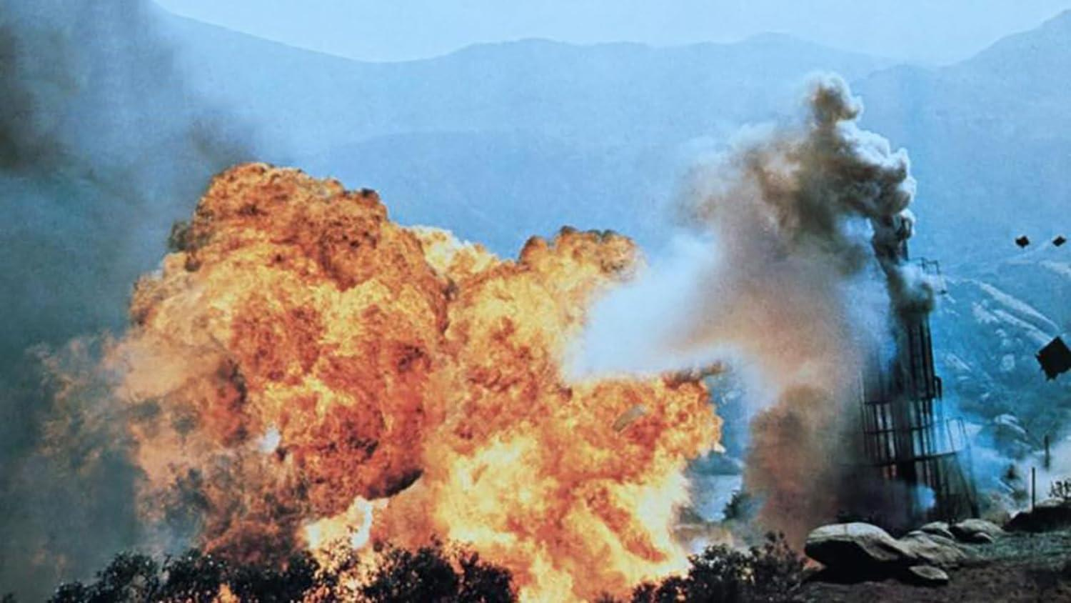 Eine Explosion bricht aus, in der Nähe eines hohen Bauwerks, mit aufsteigendem Rauch. Der Himmel ist blau und Berge sind im Hintergrund. Feuer verschlingt die Szene.