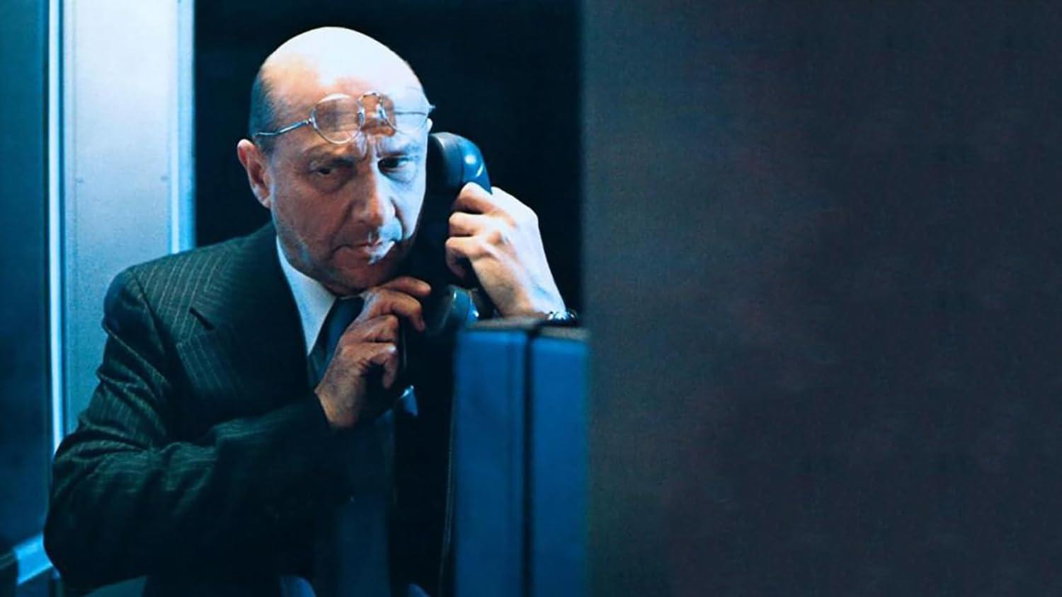 Ein Mann im Anzug hält ein Telefon an sein Ohr. Er hat eine Brille auf der Stirn. Dunkler Hintergrund und blaues Telefon.