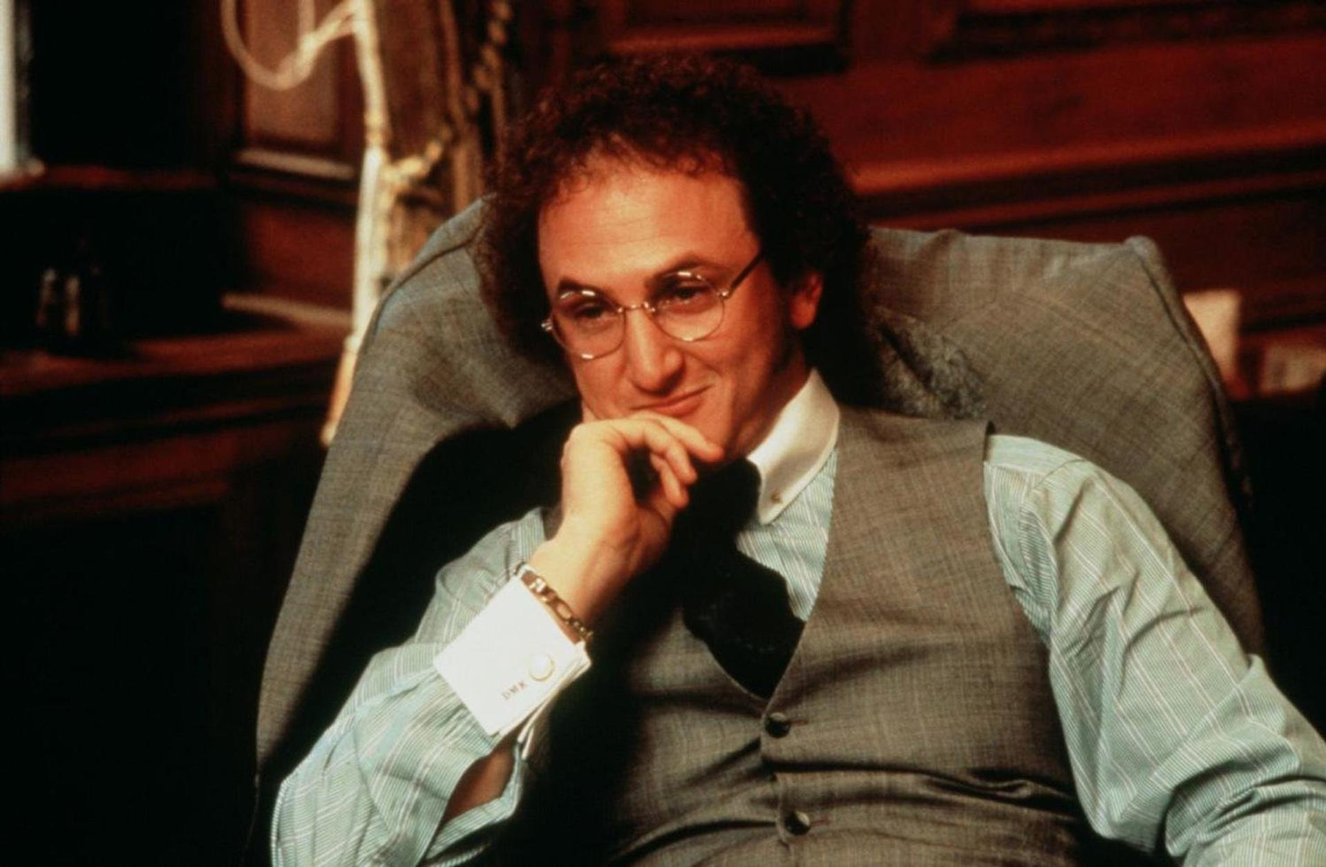 Ein Mann mit lockigem Haar und Brille sitzt in einem Stuhl, trägt eine Weste und ein Hemd und stützt sein Kinn auf seine Hand.