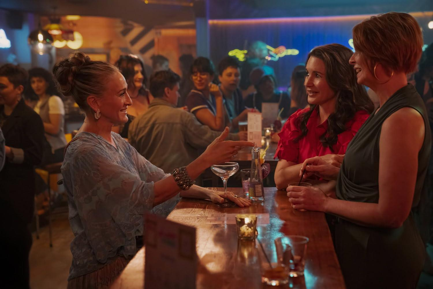 Drei Frauen an einer Bar, die sich unterhalten. Getränke und Gläser stehen auf der Theke. Im Hintergrund sind Menschen, über der Bar Neonlichter.