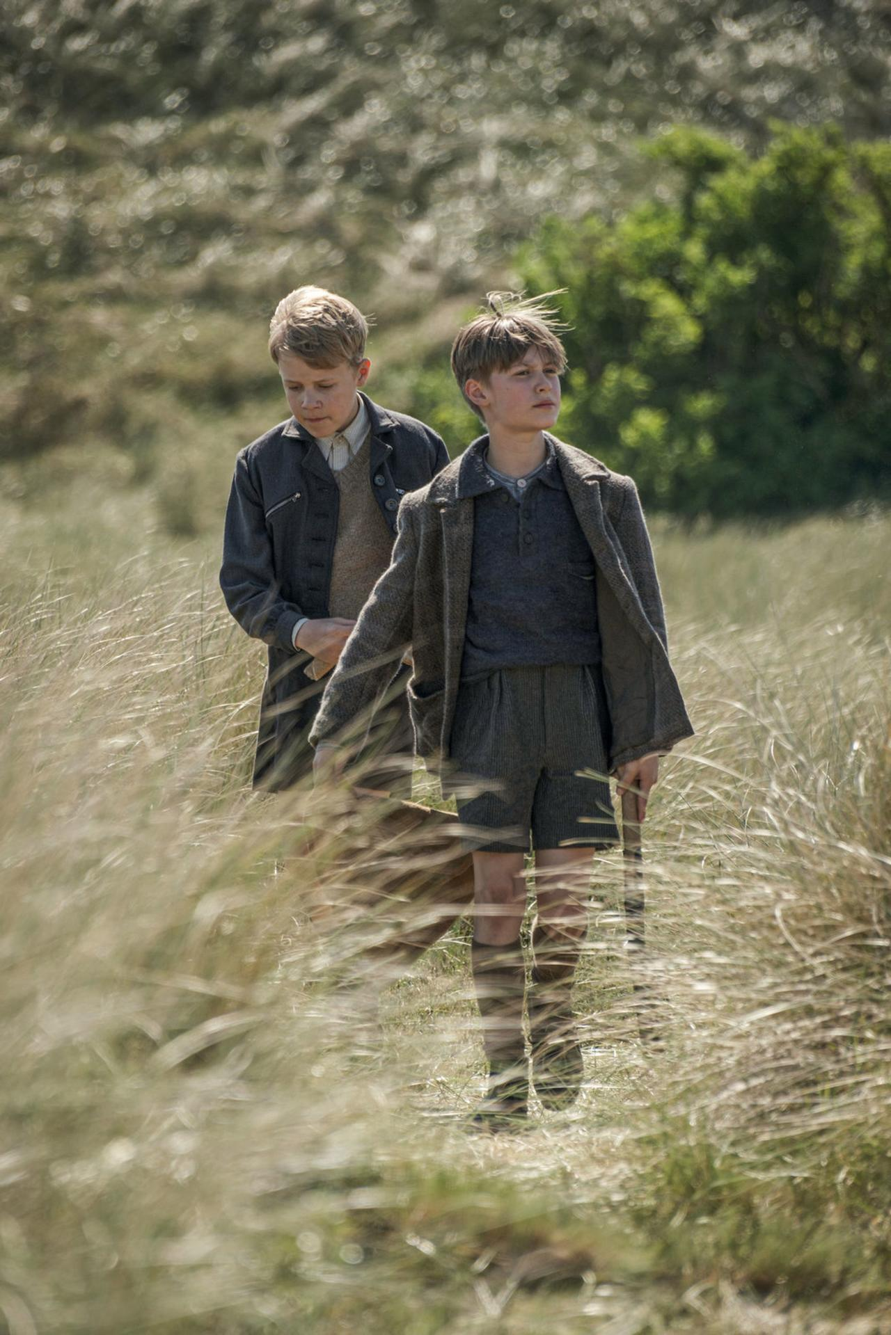 Zwei junge Jungen gehen durch hohes Gras. Einer trägt Shorts und eine Jacke, der andere eine Jacke und einen Pullover. Sonnenlicht filtert hindurch.