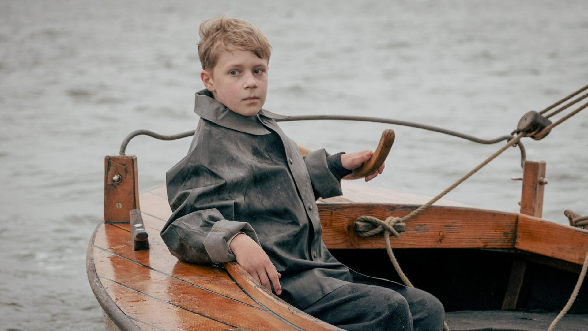 Ein Junge in einem grauen Mantel sitzt in einem Holzboot und hält eine hölzerne Pinne. Der Hintergrund ist ein graues Meer.