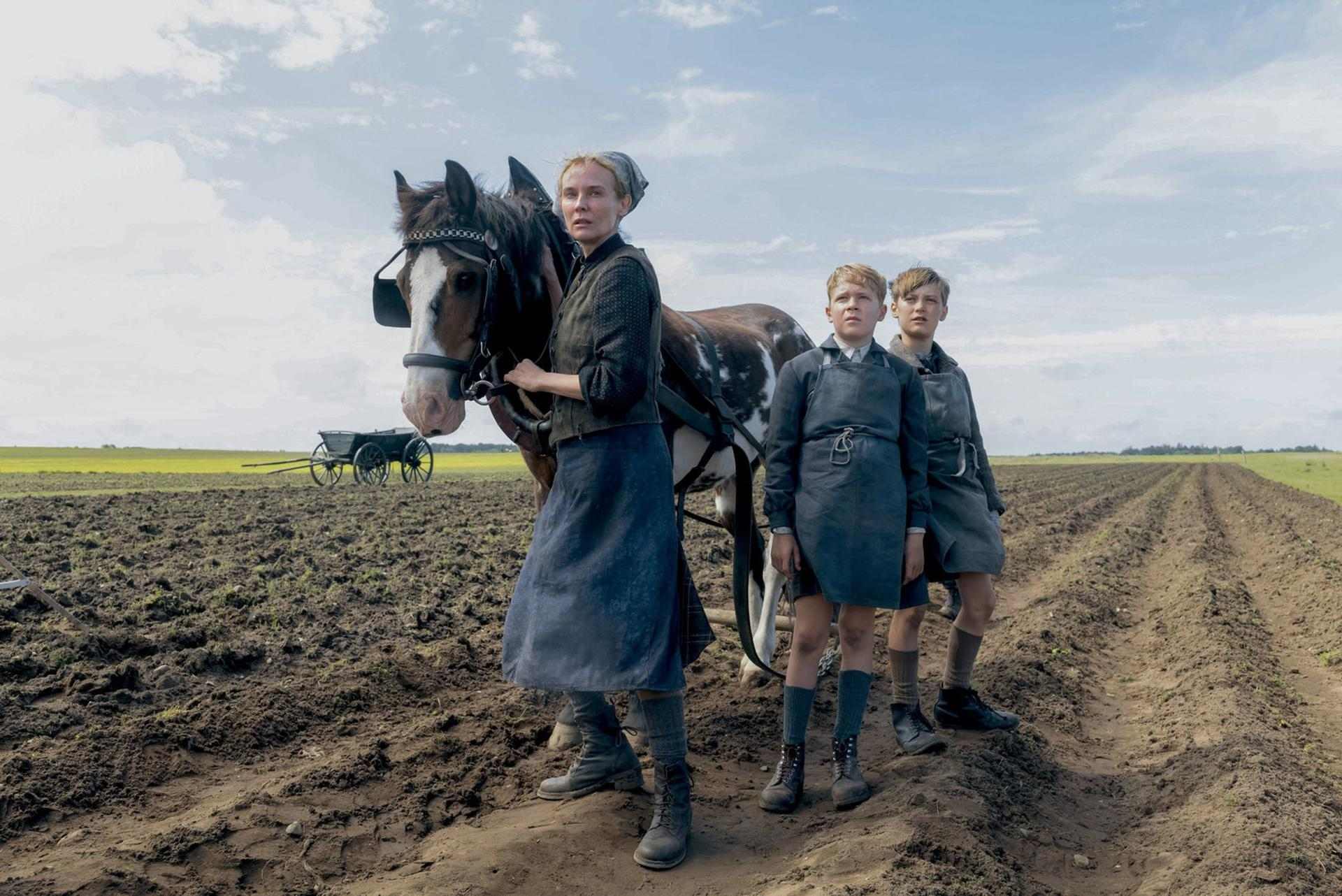 Eine Frau und zwei Jungen stehen mit einem Pferd auf einem Feld. Das Pferd ist an einen Wagen angespannt. Der Himmel ist teilweise bewölkt.