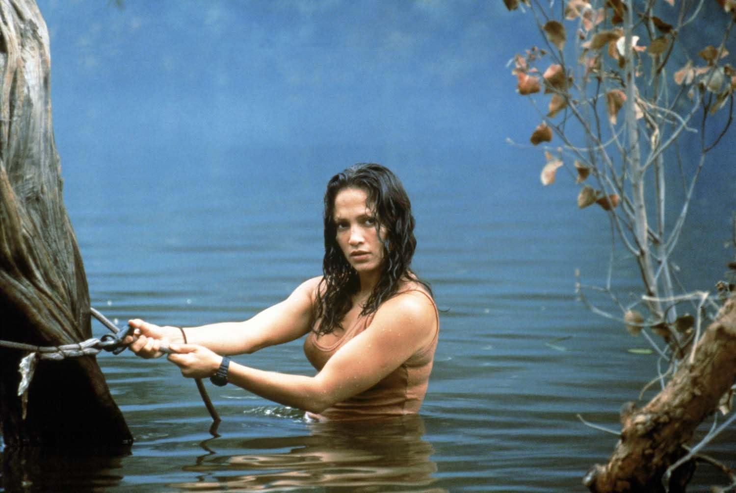 Eine Frau in einem braunen Oberteil steht im Wasser und hält ein Seil. Bäume rahmen die Szene ein, im Hintergrund ein blauer See.