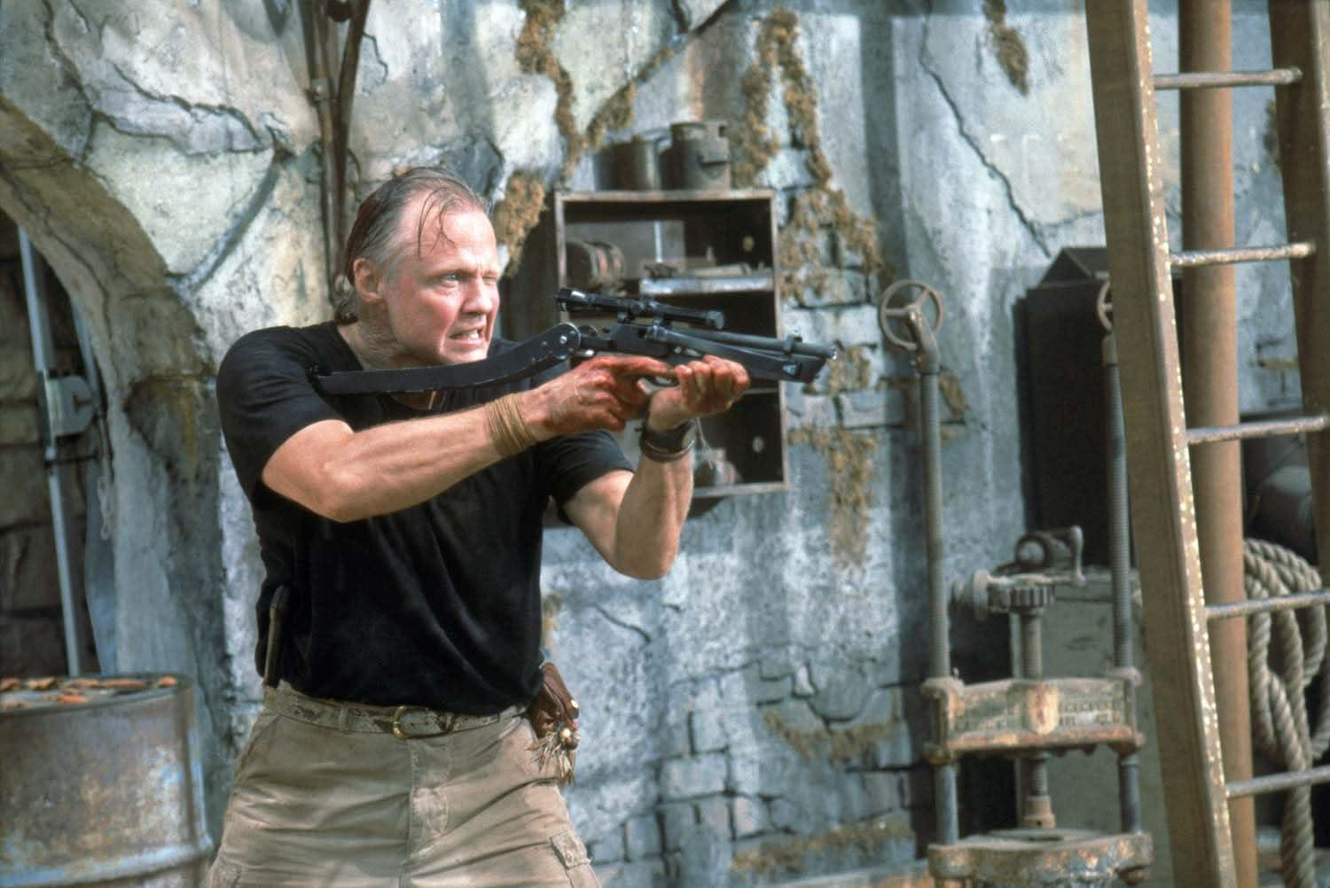 Mann zielt mit einem Gewehr in einem Steinraum. Er trägt ein schwarzes Hemd und khakifarbene Hosen. Die Szene hat industrielle Elemente.