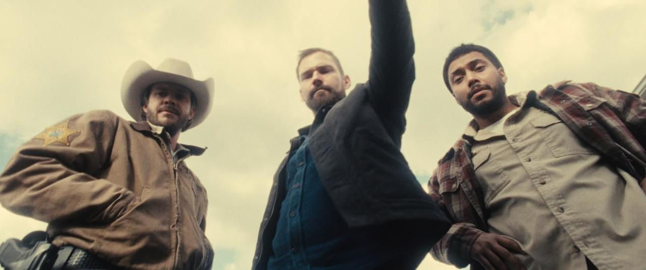Drei Männer schauen den Betrachter an. Einer trägt einen Cowboyhut, ein anderer eine Jacke und der letzte ein kariertes Hemd.