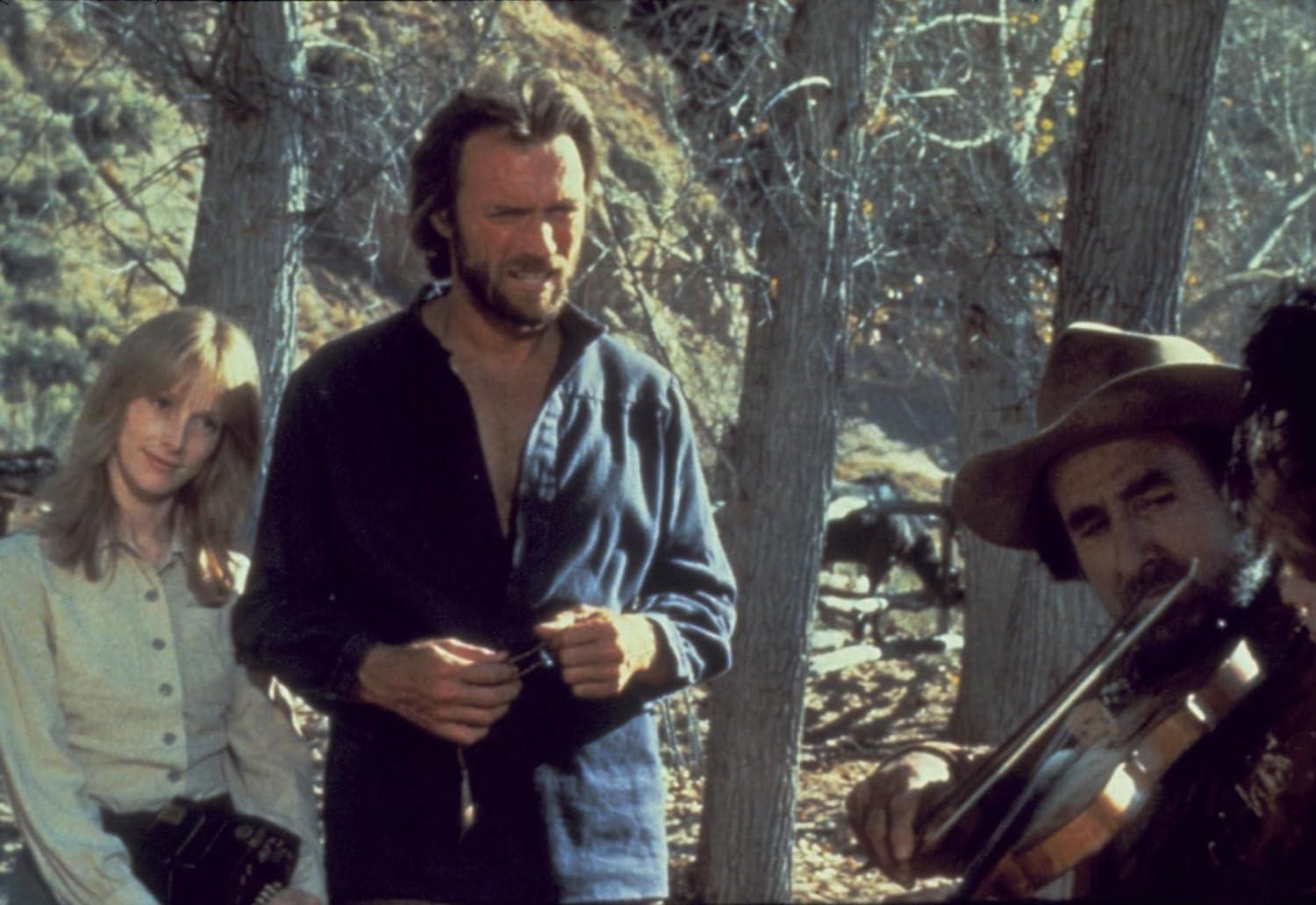 Clint Eastwood mit einer Frau und einem Geiger in einer Waldszene. Eastwood hält einen Gegenstand. Ein Mann spielt die Geige.
