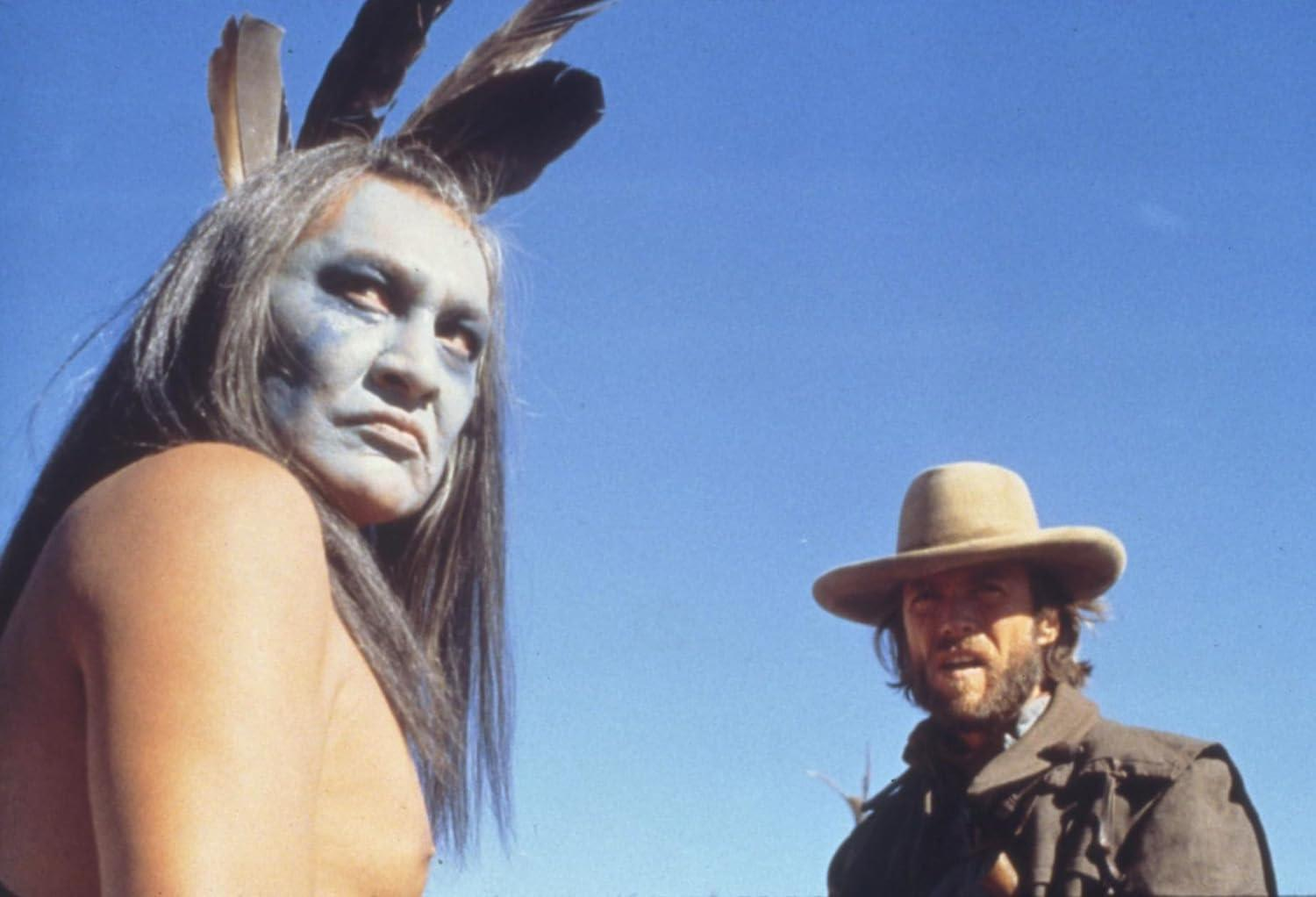 Indianer mit blauer Gesichtsfarbe und Federn und ein Mann mit Hut vor blauem Himmel.