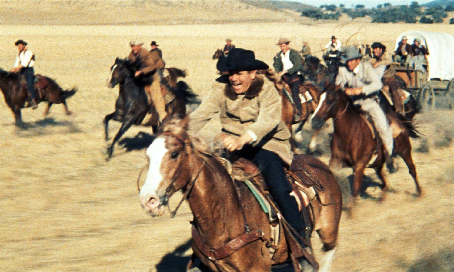 Cowboys auf Pferden galoppieren über eine staubige Ebene. Ein Planwagen ist im Hintergrund zu sehen, ebenso wie sanfte Hügel.