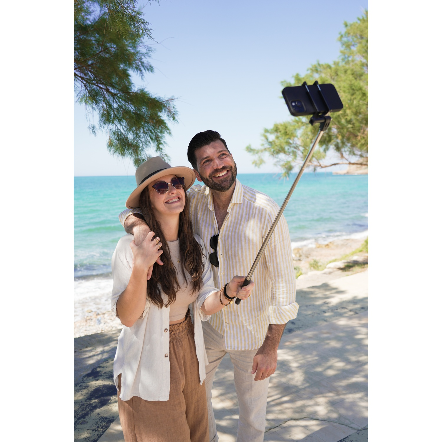 Una coppia si fa un selfie in spiaggia. La donna indossa un cappello e occhiali da sole, l'uomo tiene l'asta per selfie. Il mare è sullo sfondo.