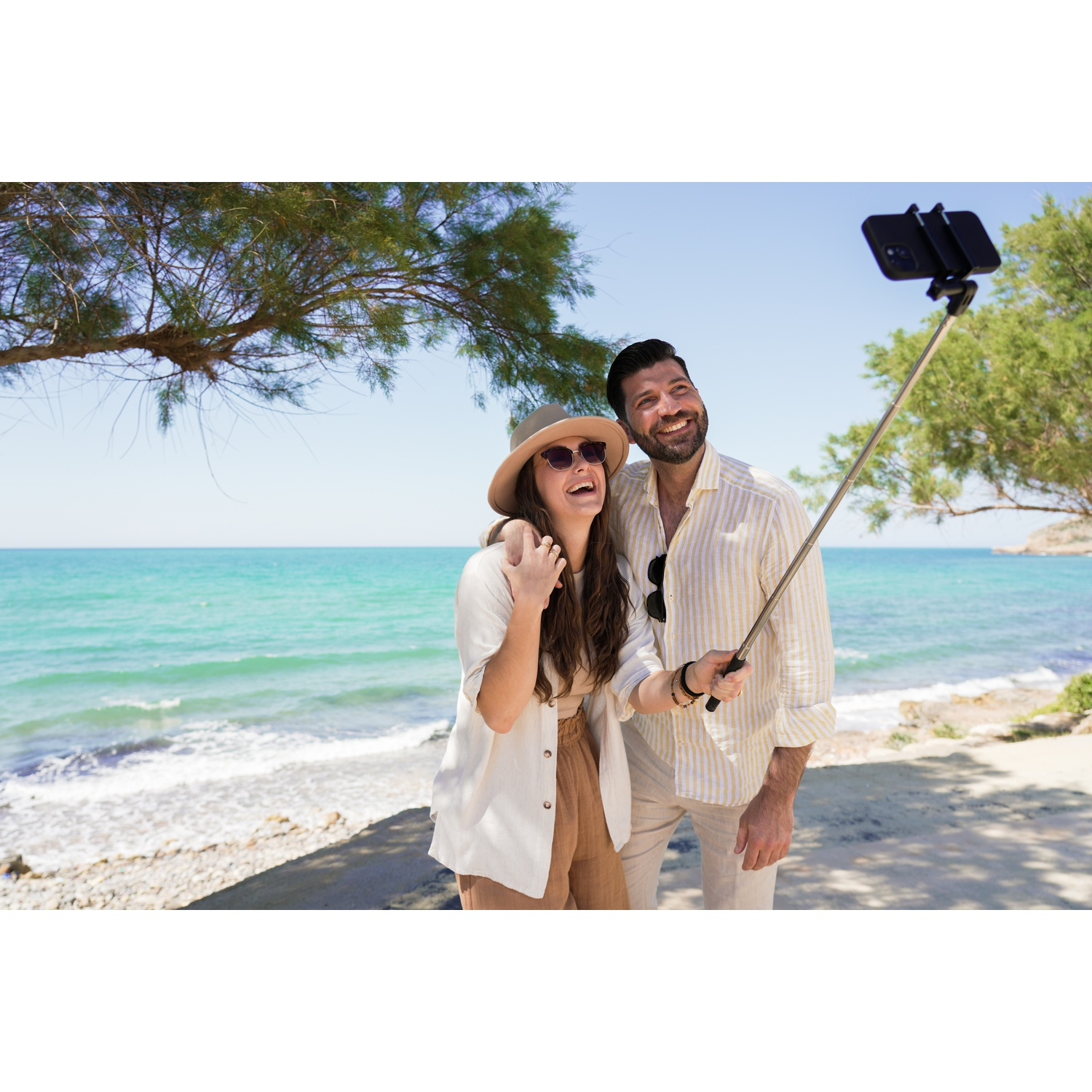Una coppia scatta un selfie sulla spiaggia. La donna indossa un cappello. L'uomo tiene il bastone per selfie. Mare blu e alberi sullo sfondo.