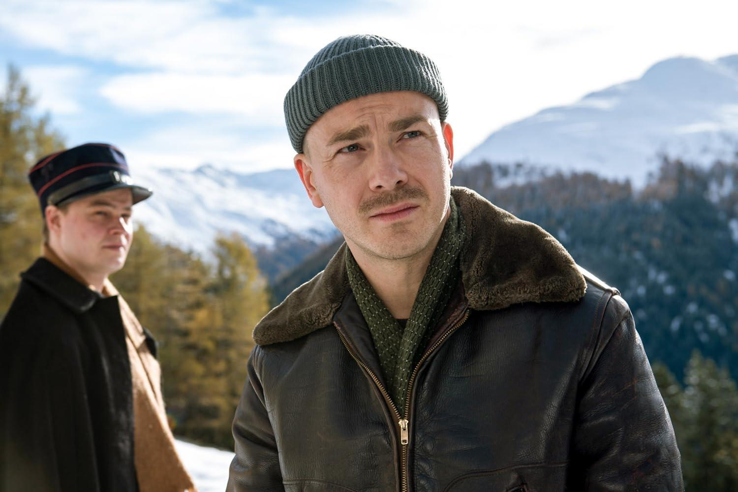 Mann mit Hut und Lederjacke blickt weg. Ein anderer Mann in Uniform steht im Hintergrund, schneebedeckte Berge.