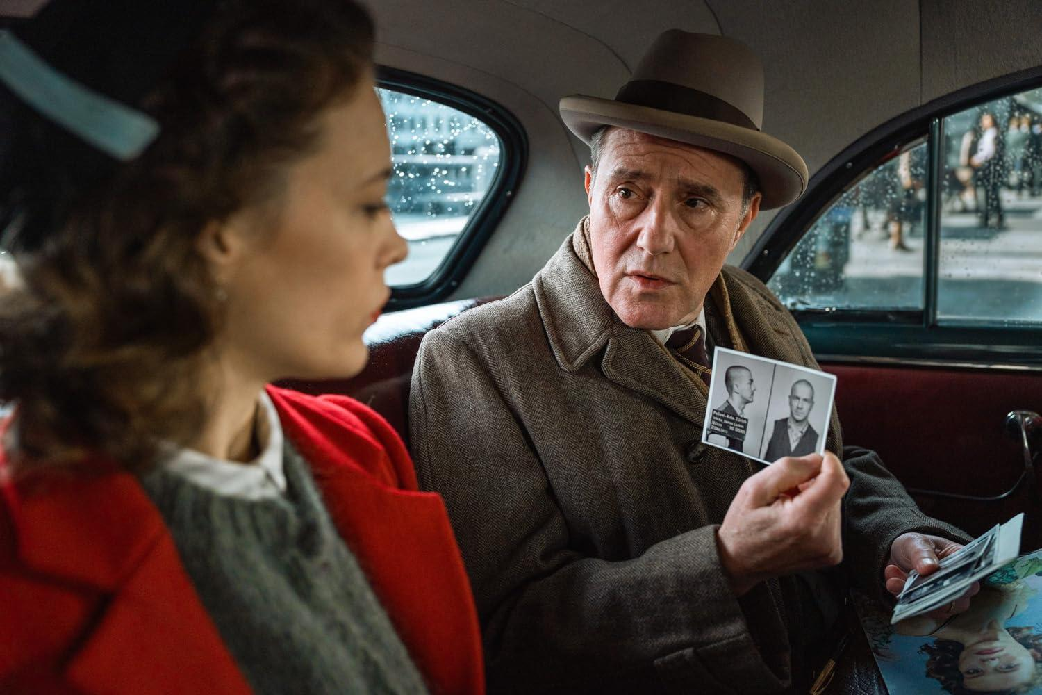 In einem Auto zeigt ein Mann mit Hut einer Frau in Rot ein Foto. Das Foto zeigt zwei Männer. Straßenszene durch das Fenster sichtbar.