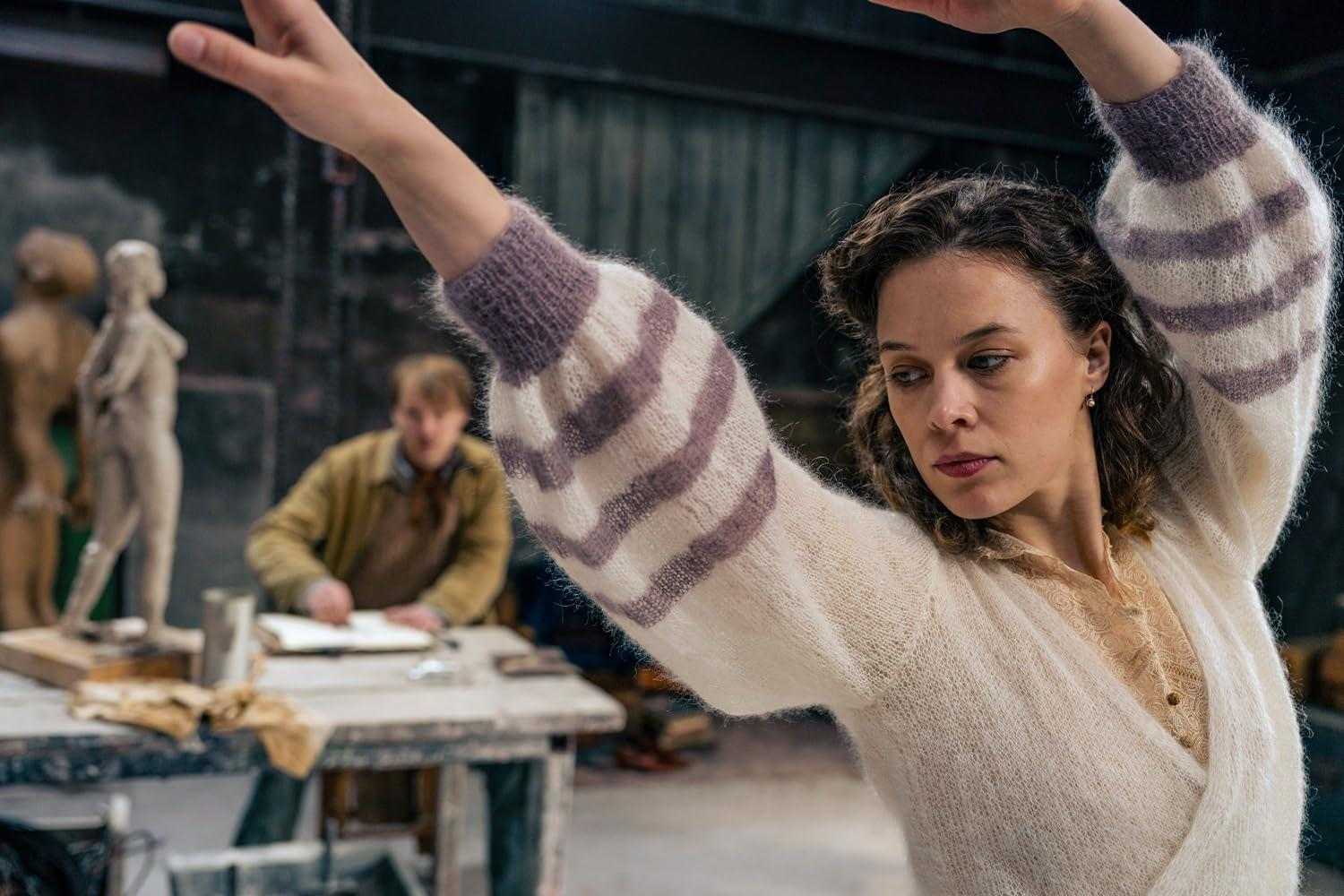Frau im Studio mit erhobenen Armen, trägt einen weißen Pullover mit lila Streifen. Mann im Hintergrund, arbeitet. Skulpturen links.