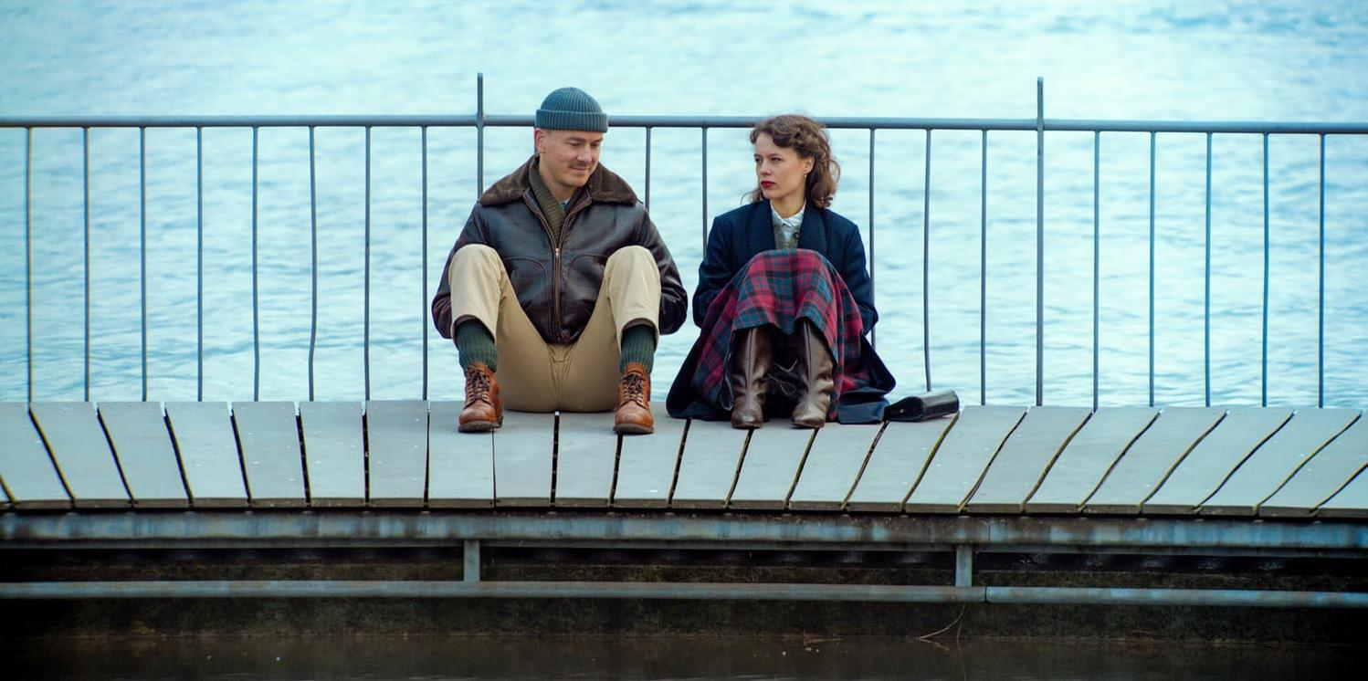 Zwei Personen sitzen auf einem Holzsteg am Wasser. Mann trägt Hut und Jacke. Frau trägt Rock und Mantel.