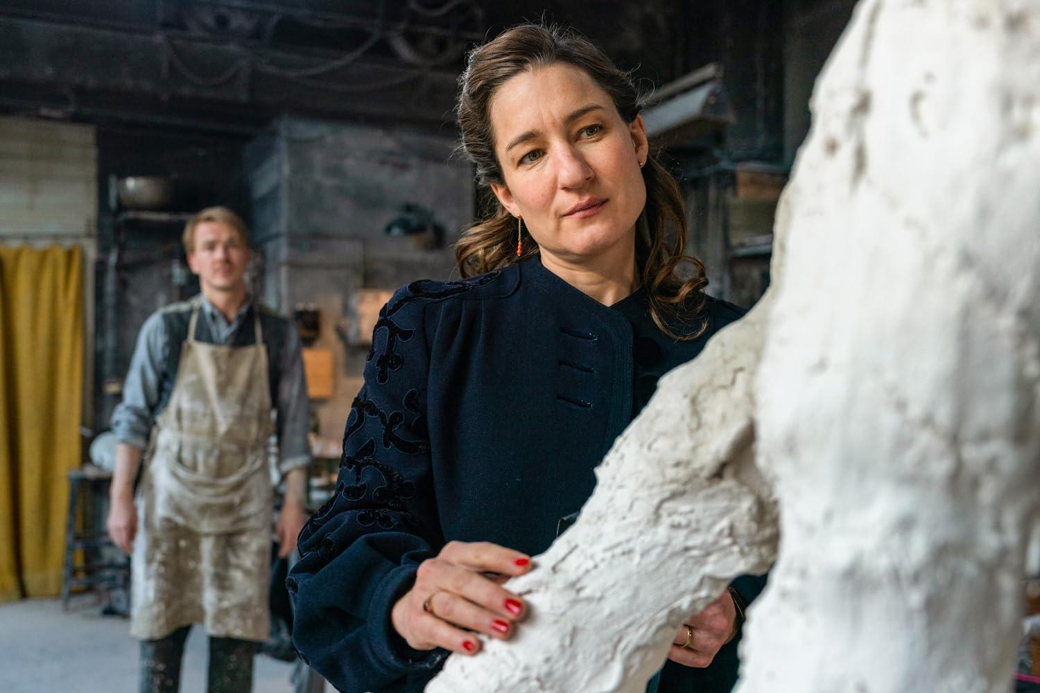 Eine Frau in einem dunklen Mantel mit roten Nägeln posiert neben einer weißen Skulptur. Im Hintergrund steht ein Mann mit Schürze.