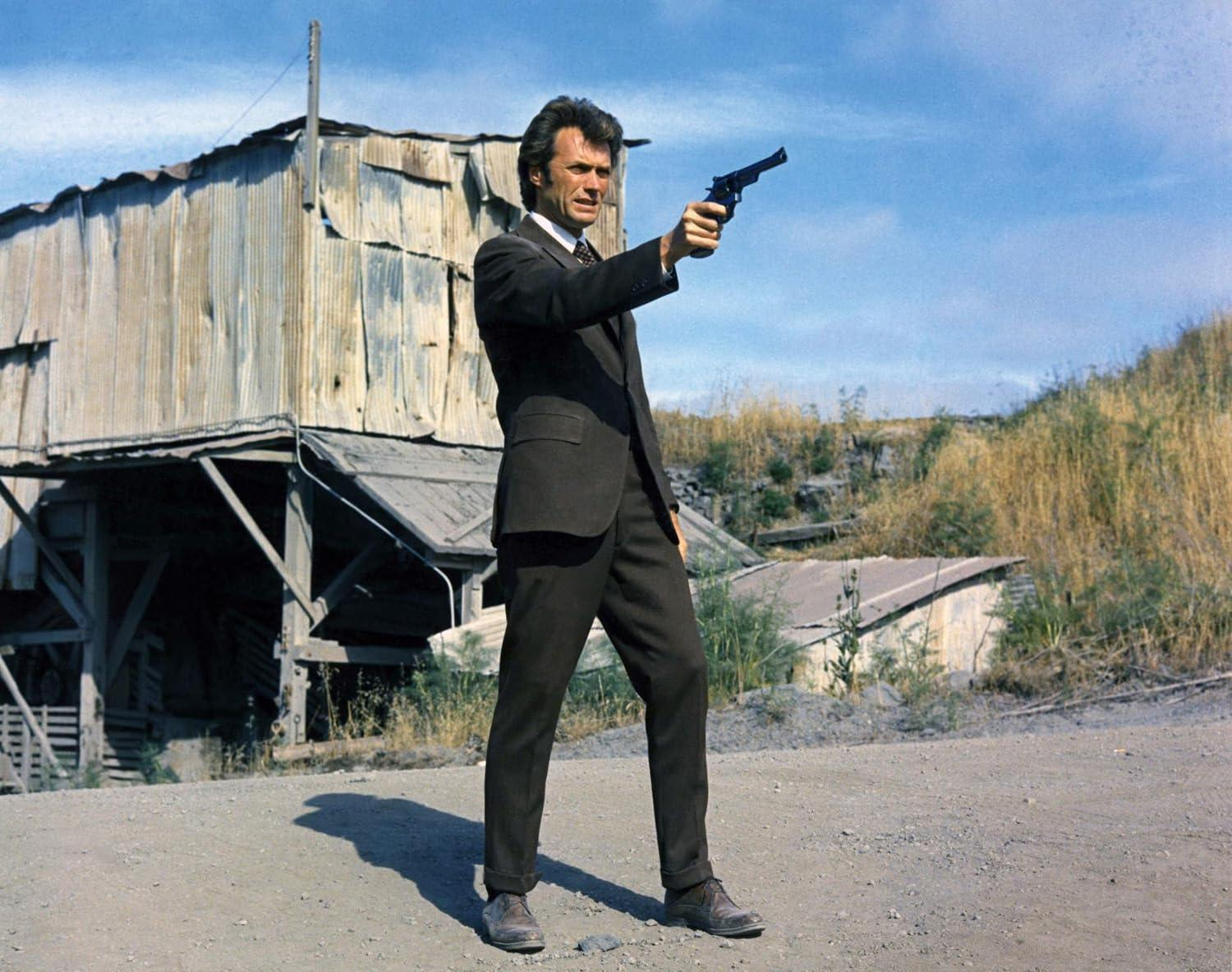 Clint Eastwood im Anzug, der eine Waffe hält. Ein verwittertes Gebäude befindet sich hinter ihm, und trockene Vegetation im Hintergrund.