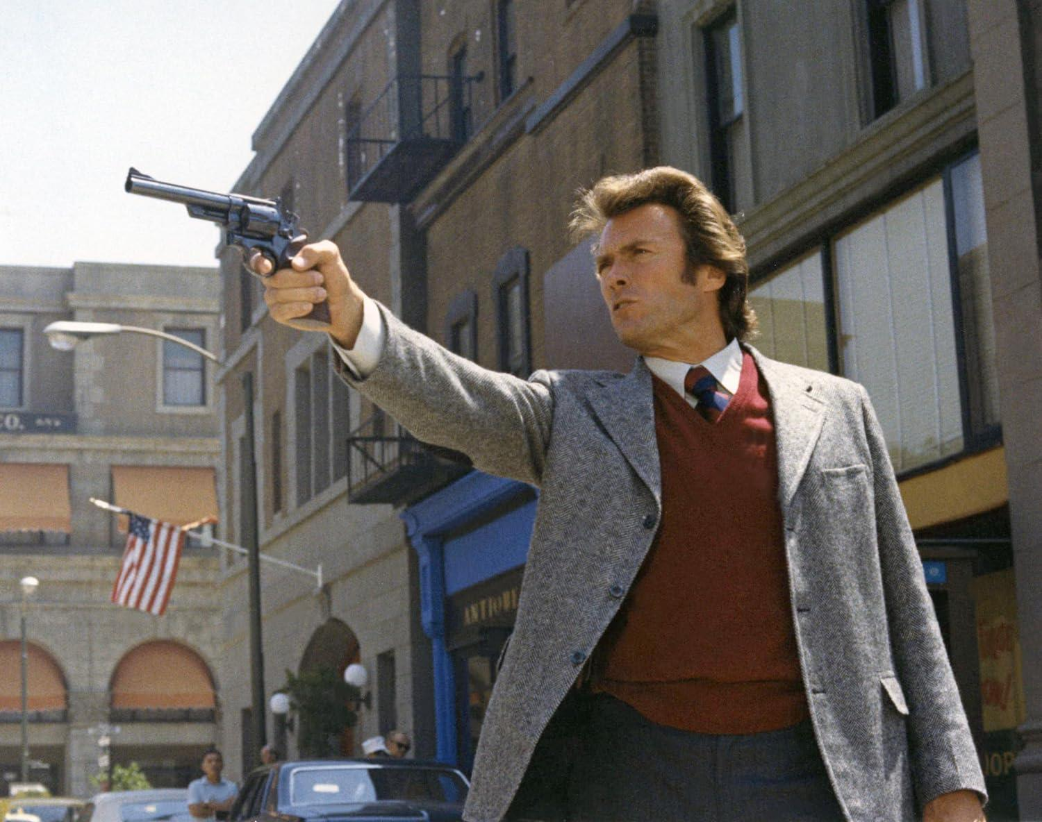 Clint Eastwood zielt mit einem Revolver. Er trägt eine graue Jacke, einen roten Pullover und eine Krawatte. Gebäude, US-Flagge und Autos im Hintergrund.