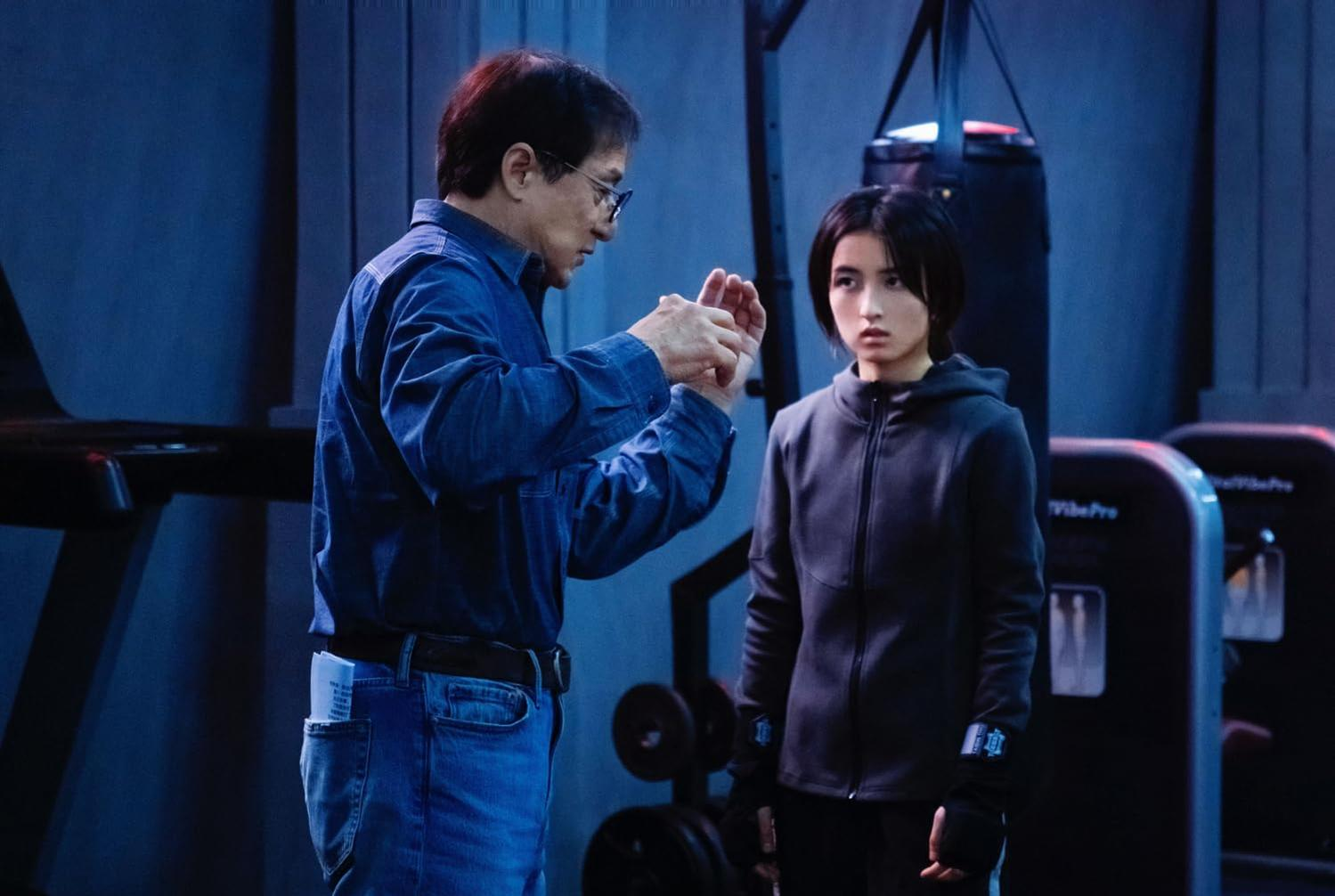 Jackie Chan gestikuliert auf eine junge Frau. Sie trägt eine Kapuzenjacke. Fitnessgeräte und ein Boxsack sind im Hintergrund.