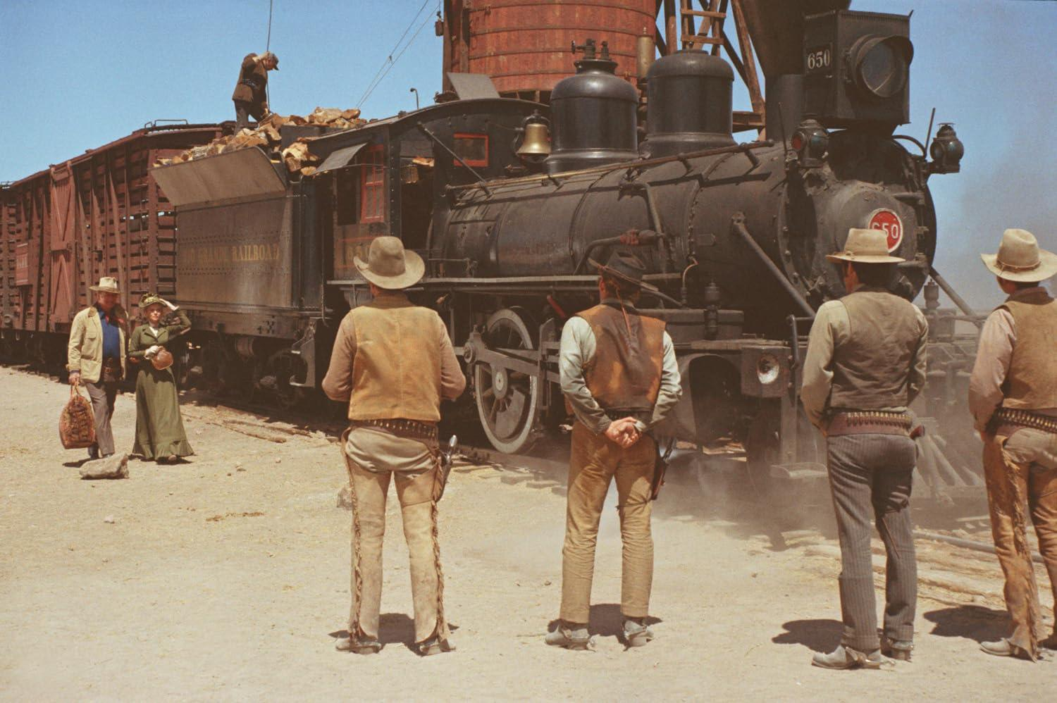 Cowboys stehen in der Nähe eines Zuges. Im Hintergrund ist eine Dampflokomotive zu sehen. Eine Frau und ein Mann stehen in der Nähe des Güterwagens des Zuges.