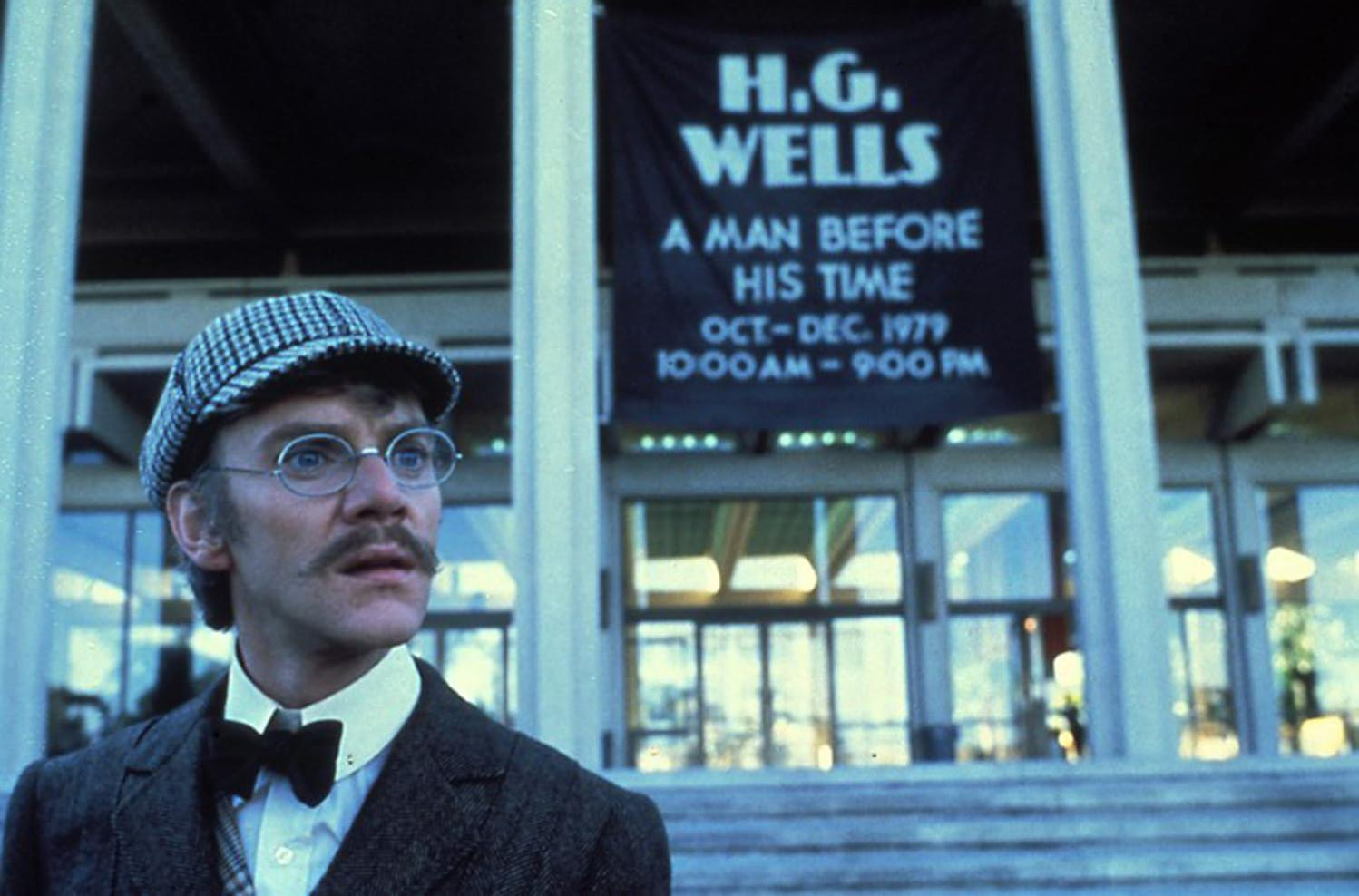 Ein Mann mit Brille und Tweedmütze steht vor einem Banner für H.G. Wells. Das Banner wirbt für eine Veranstaltung.