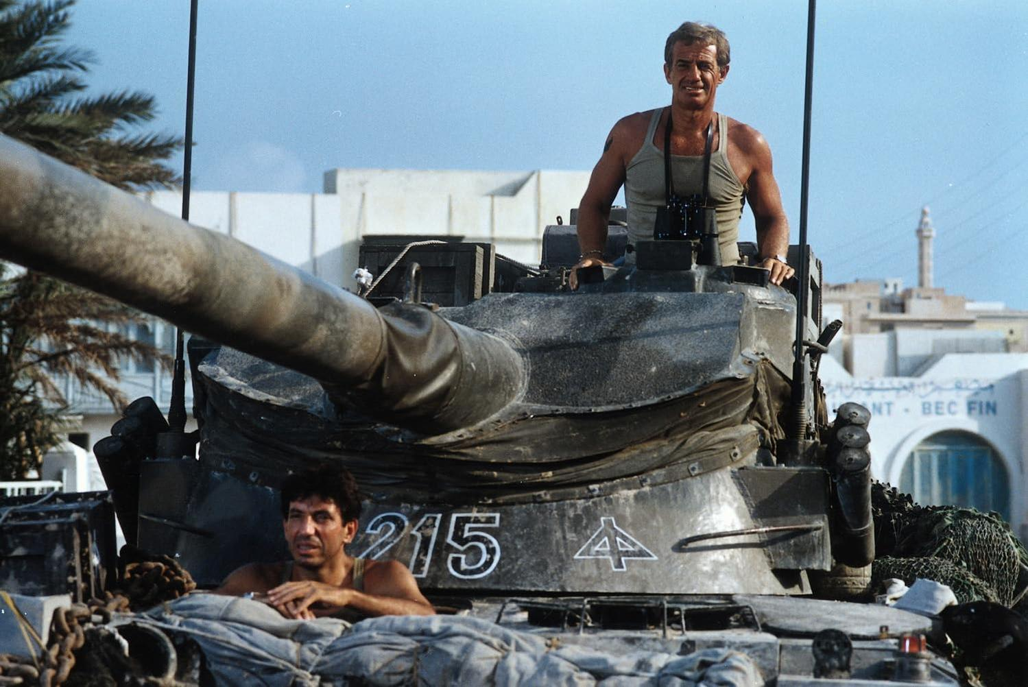 Zwei Männer auf einem Panzer. Einer steht, trägt ein Tanktop und hält ein Fernglas. Der andere sitzt tief. Gebäude und Palmen im Hintergrund.