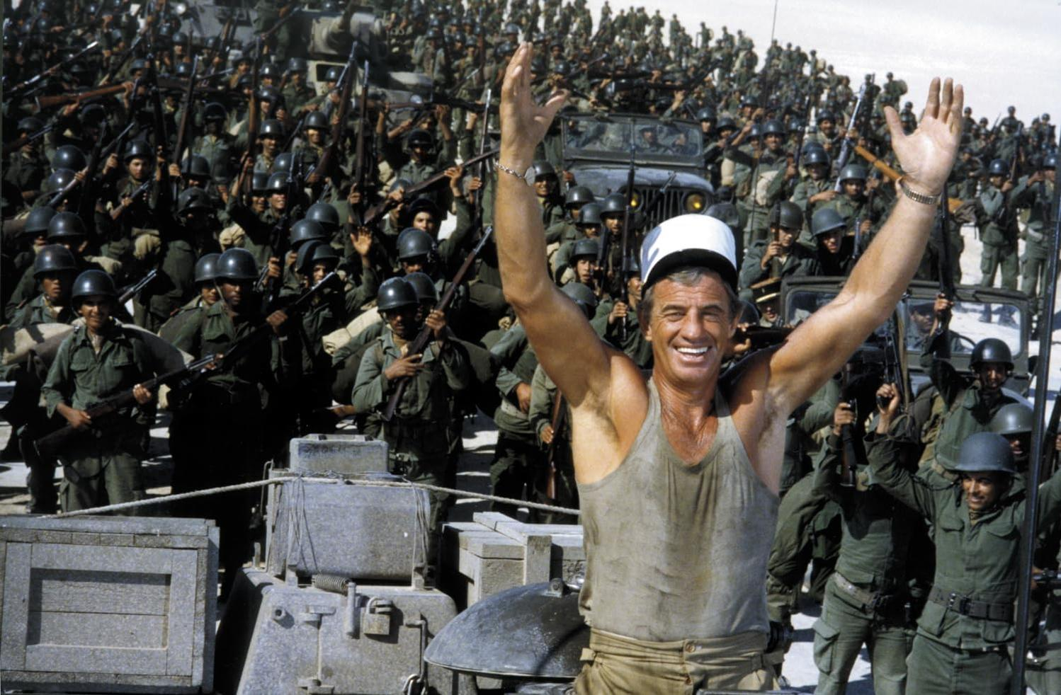 Jean-Paul Belmondo mit weißem Hut lächelt und hebt die Arme. Soldaten mit Gewehren und Helmen umgeben einen Jeep.