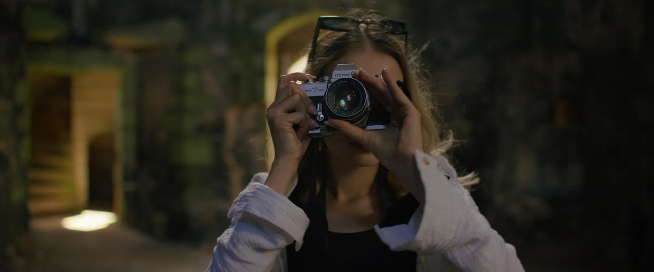 Eine Frau hält eine Kamera und macht ein Foto. Eine Sonnenbrille sitzt auf ihrem Kopf. Dunkler Steinbogen im Hintergrund.