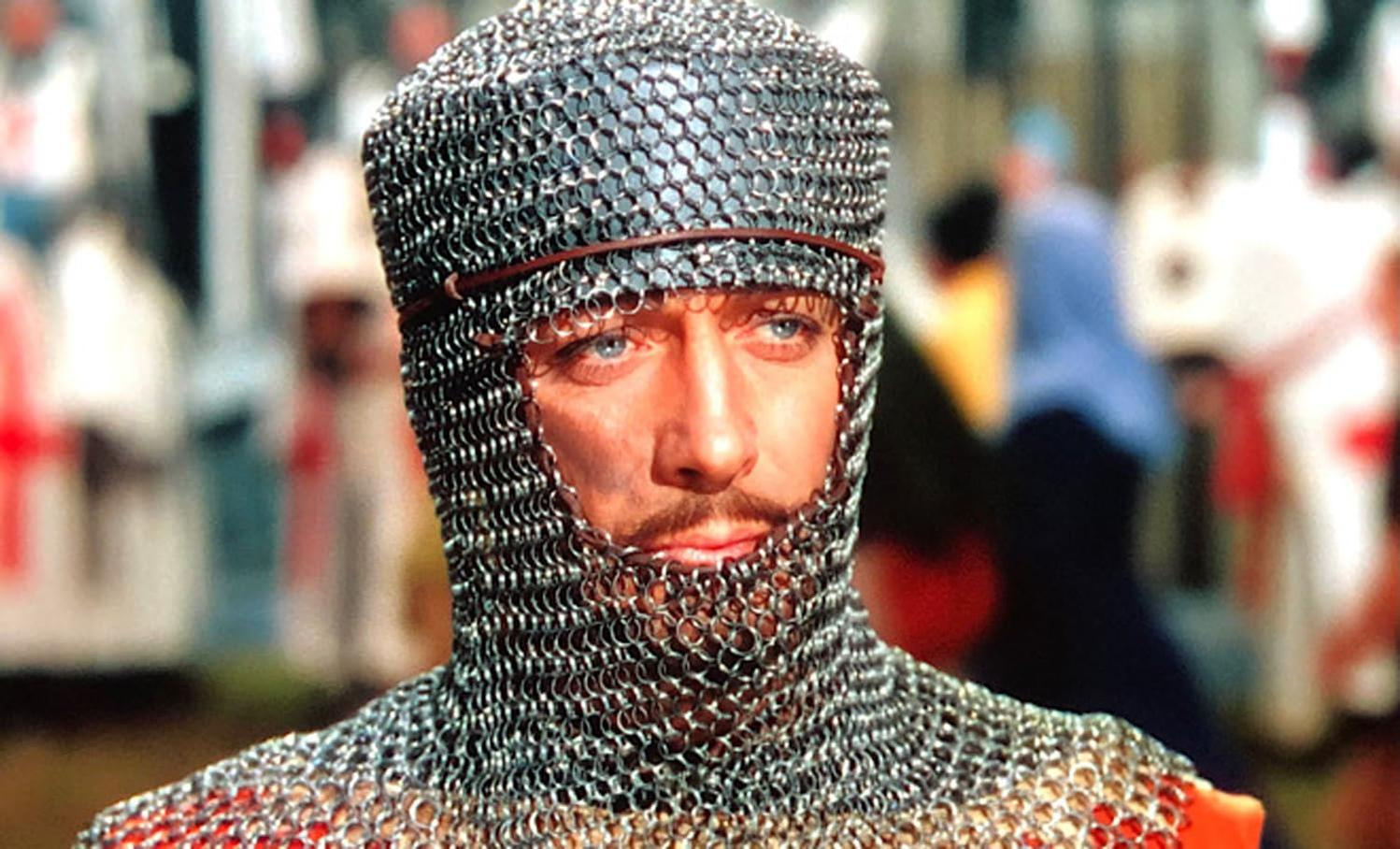 Ein Mann in Kettenrüstung und Helm blickt den Betrachter an. Im Hintergrund verschwommene Figuren.