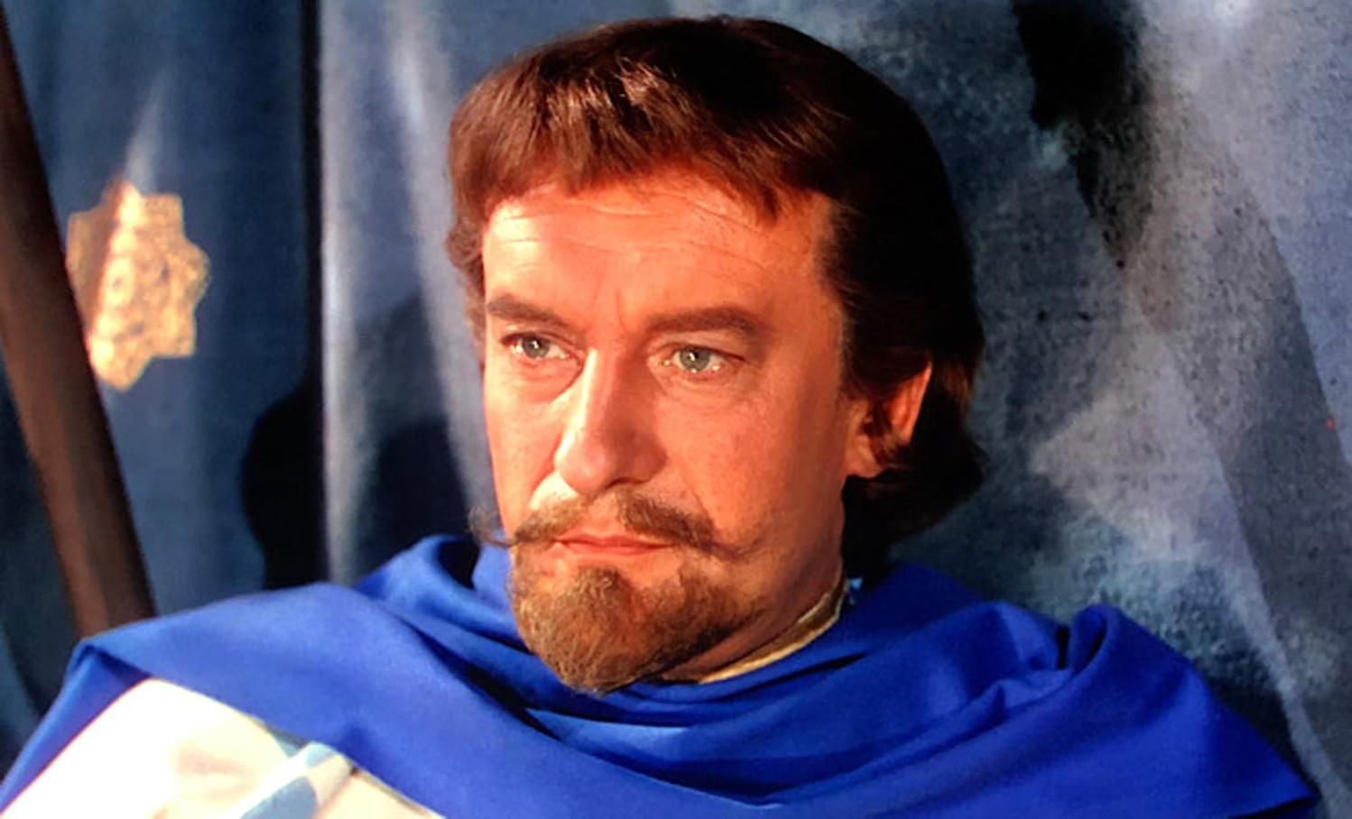 Ein Mann mit braunem Haar und Bart, der einen blauen Umhang trägt, blickt nach links. Der Hintergrund ist ein blauer Vorhang mit einem Stern.
