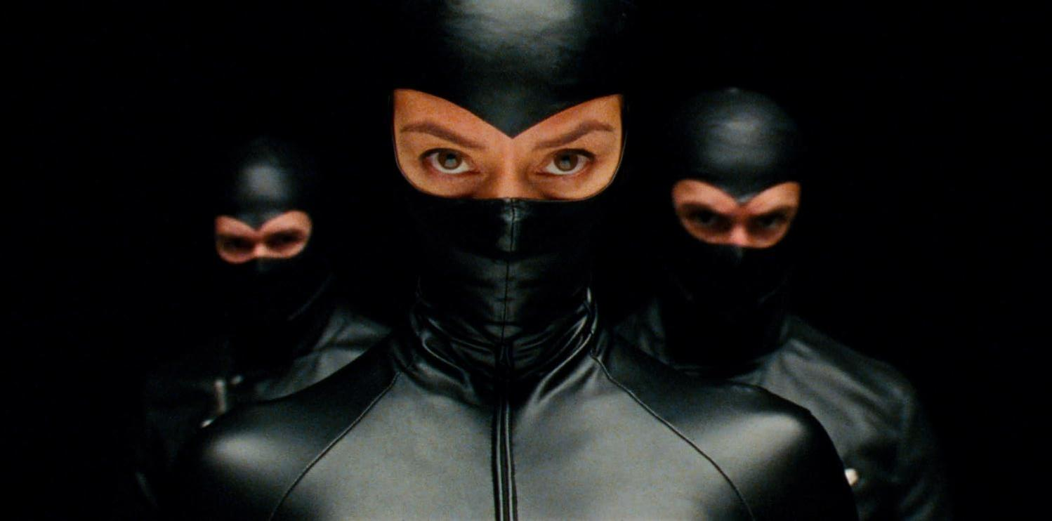 Drei Personen mit schwarzen Masken und Outfits sind vor einem schwarzen Hintergrund zentriert. Nur ihre Augen sind sichtbar.