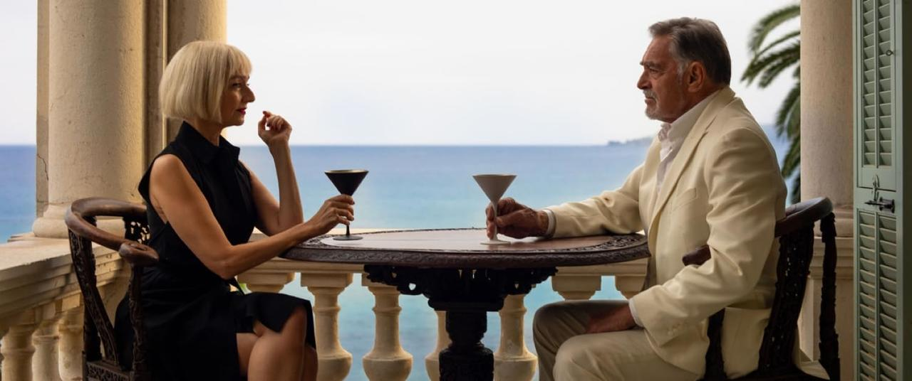 Ein Mann und eine Frau sitzen an einem Tisch und halten Cocktails. Sie befinden sich auf einem Balkon mit Blick auf das Meer. Palmen und Fensterläden sind zu sehen.