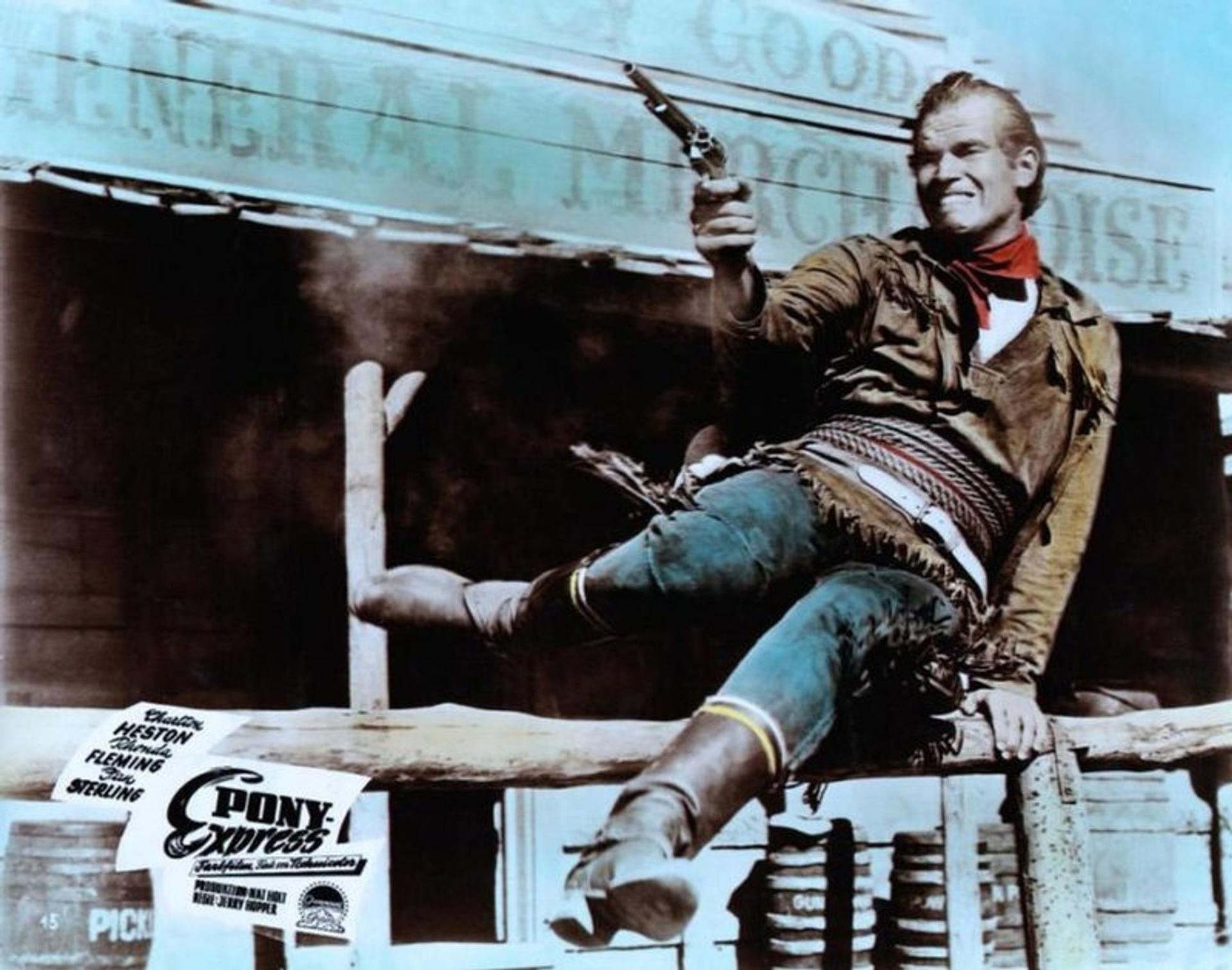 Ein Mann in Cowboy-Kleidung zielt mit einer Waffe. Er sitzt auf einer Holzreling mit einem Schild, auf dem 'Pony Express' steht. Im Hintergrund befindet sich ein Schild.