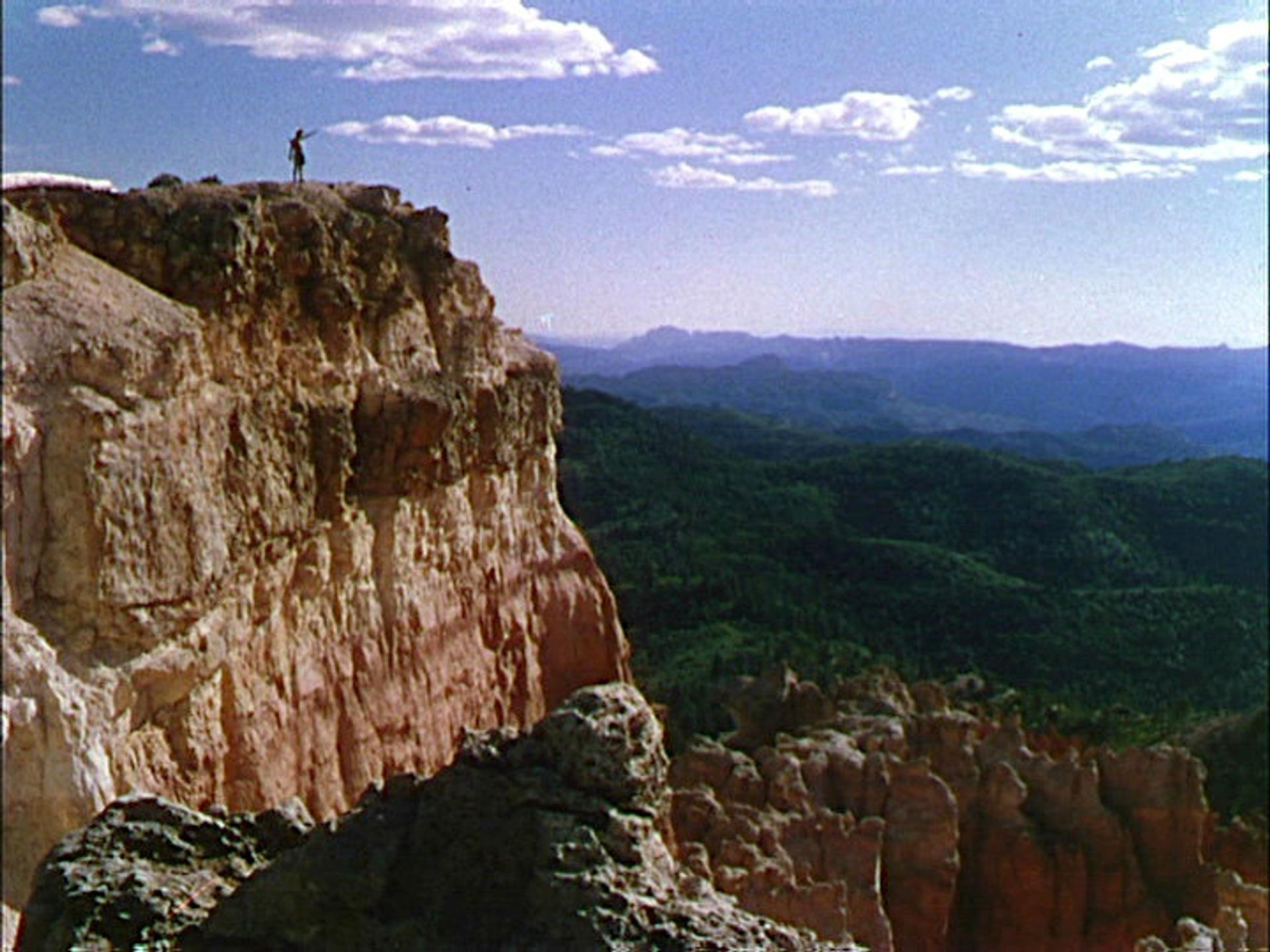 Eine Person steht auf einer Klippe und blickt auf grüne Hügel und einen blauen Himmel mit Wolken. Die Klippe ist hellbraun.