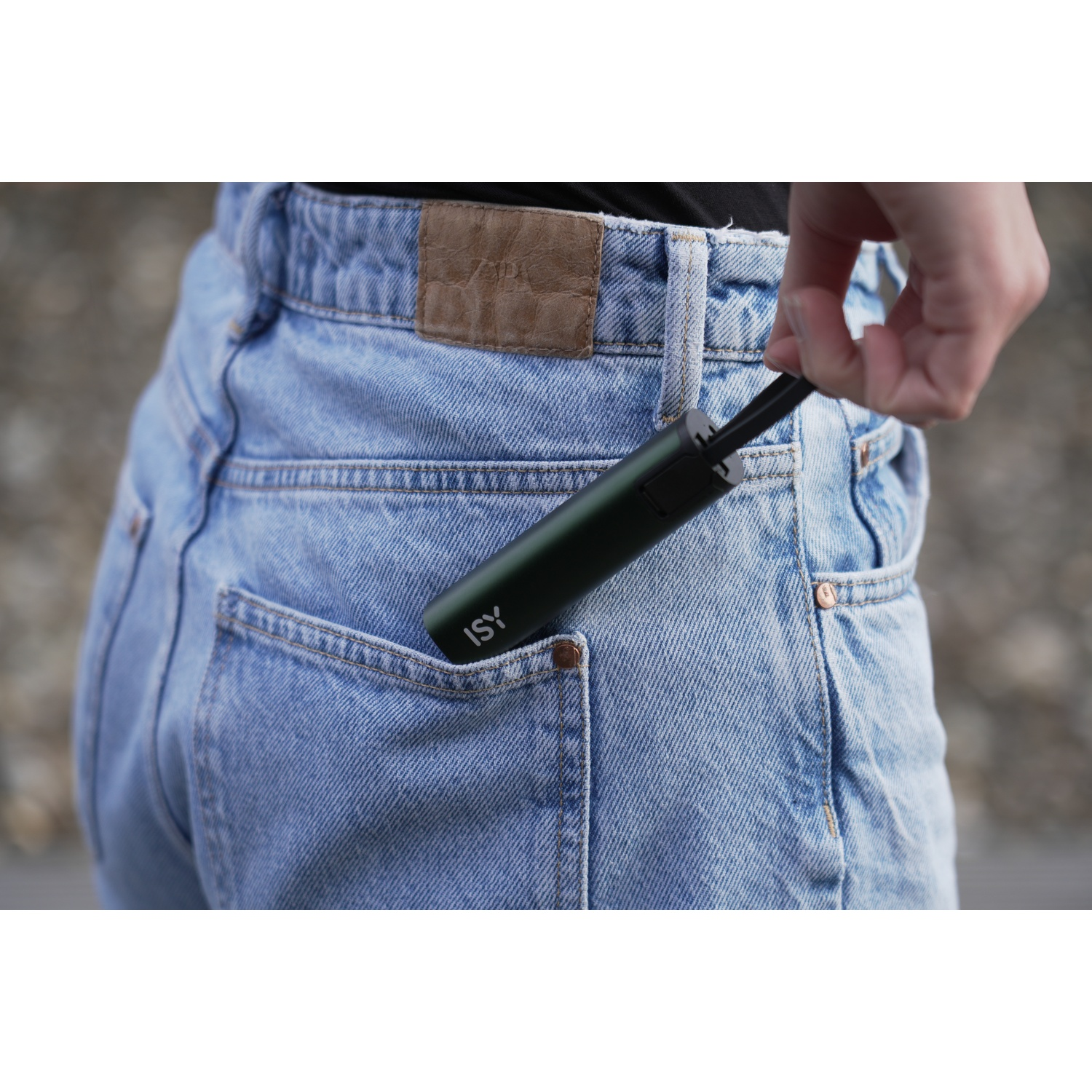 Una mano tira fuori un dispositivo ISY verde da una tasca dei jeans. I jeans sono azzurri, con una toppa in pelle marrone sul retro.