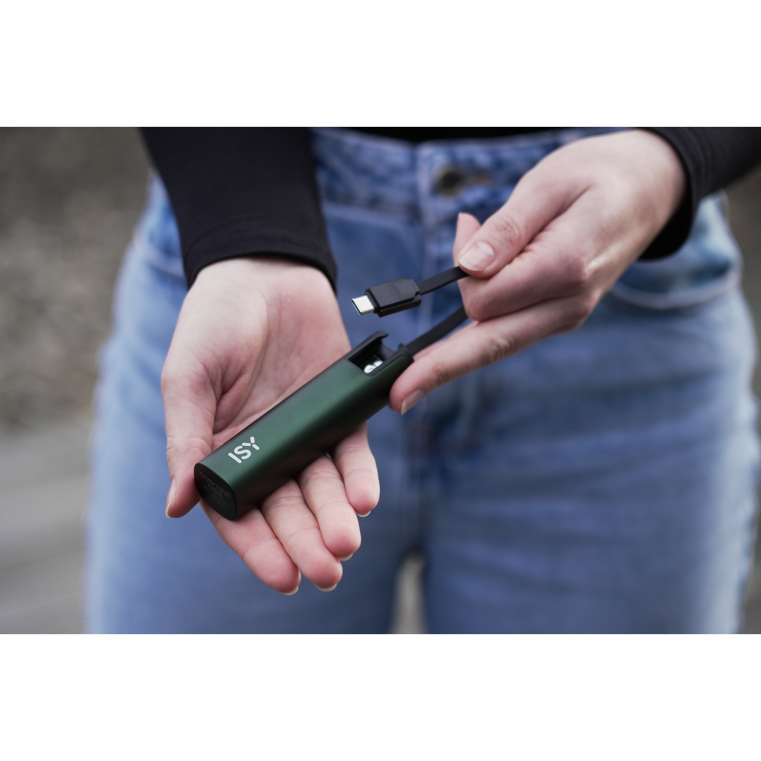Una persona tiene in mano un power bank ISY verde e un cavo USB-C. Il power bank è in una mano, il cavo nell'altra. Jeans sullo sfondo.