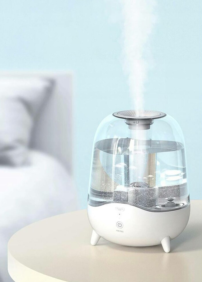Biały nawilżacz z przezroczystym zbiornikiem na wodę stoi na stole. Para unosi się w górę. Niebieskie tło.