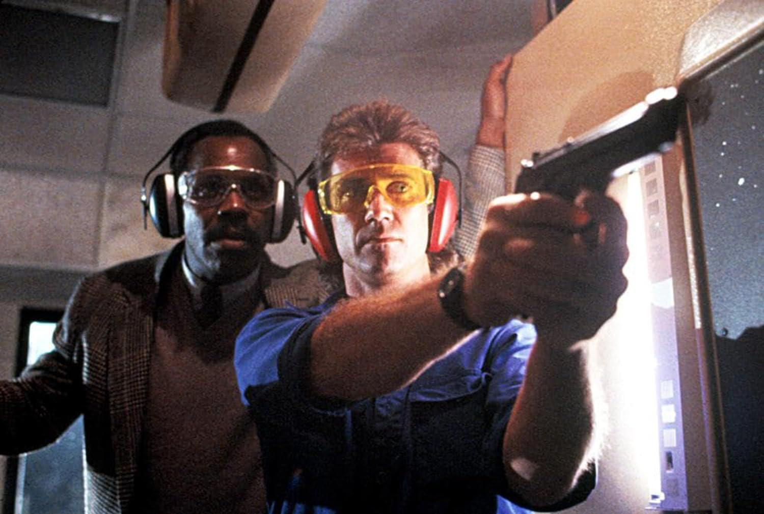 Zwei Männer in einem Schießstand. Einer zielt mit einer Waffe, trägt Ohrenschützer und Brille. Der andere beobachtet. Im Hintergrund ist ein Fenster.