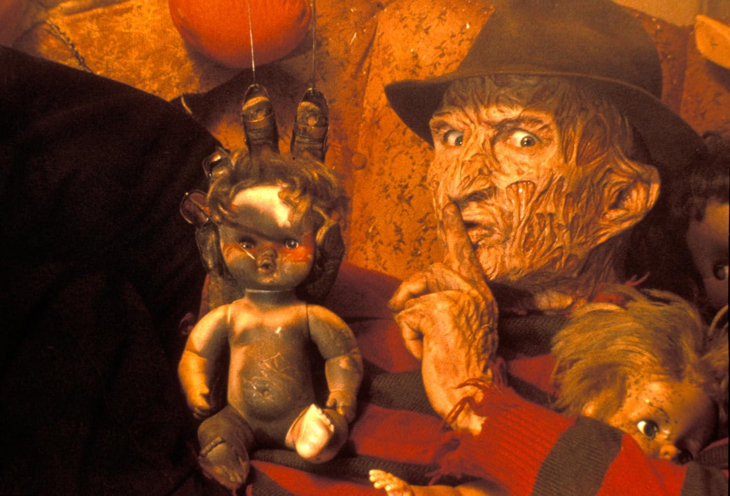 Freddy Krueger mit Puppe, Hut, Finger auf den Lippen. Die Puppe hat lockiges Haar. Rot-schwarz gestreifter Pullover.
