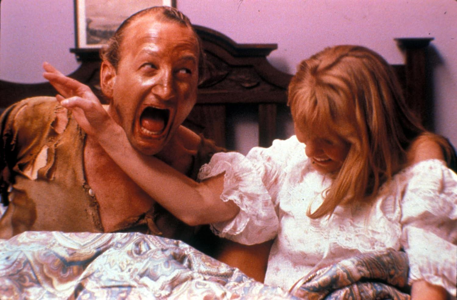 Ein Mann schreit entsetzt, Arm erhoben. Eine Frau in einem weißen Kleid ist neben ihm. Sie sind auf einem Bett. Der Hintergrund ist ein dunkles Kopfteil.