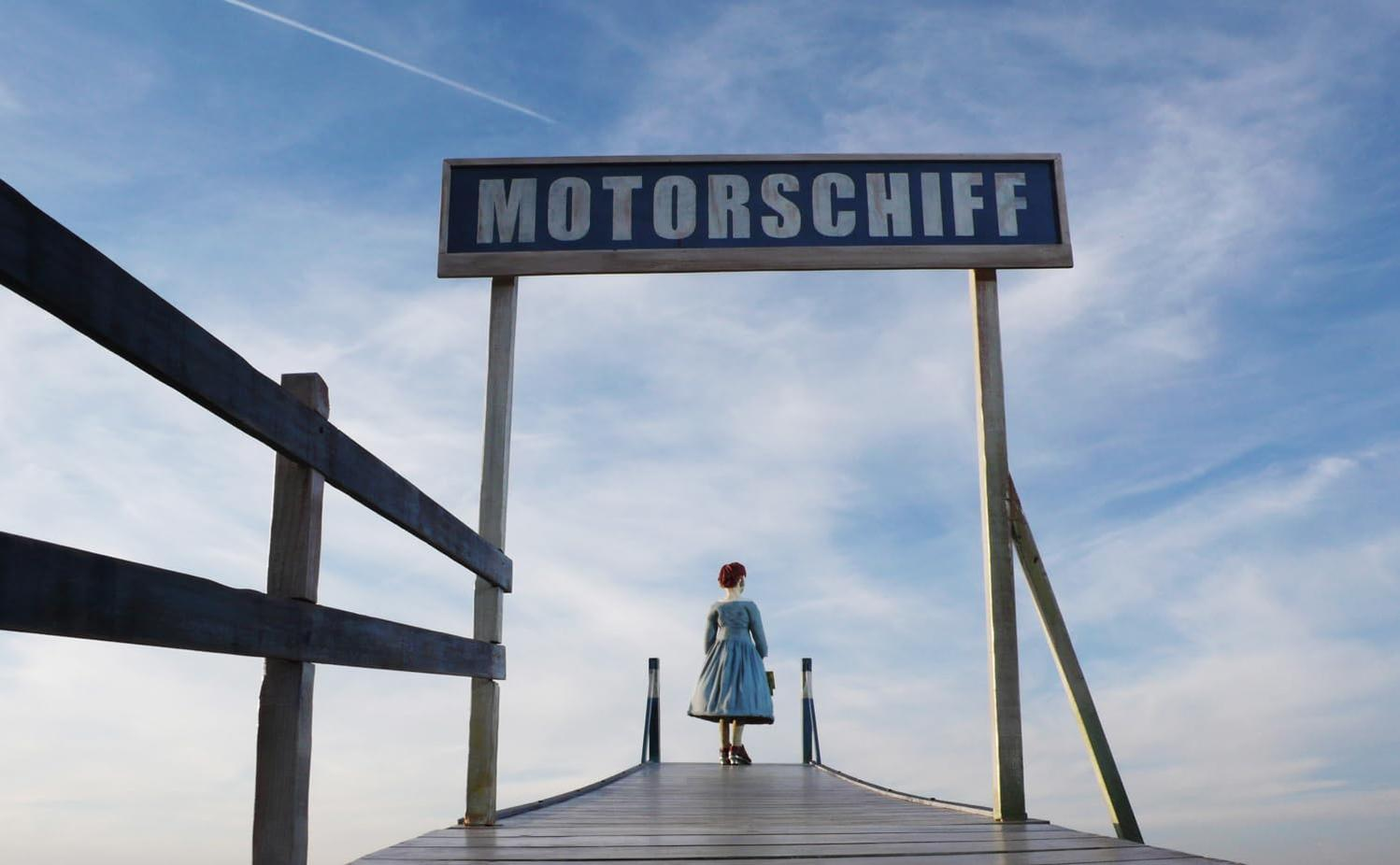 Eine Frau in einem blauen Kleid steht auf einem Holzsteg unter einem Schild mit der Aufschrift 'MOTORSCHIFF' vor blauem Himmel.