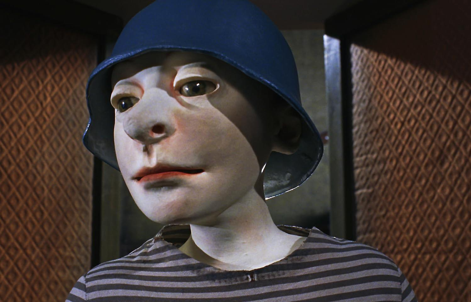 Eine Marionette mit blauem Hut und gestreiftem Hemd. Die Marionette hat ein blasses Gesicht und rote Lippen. Der Hintergrund hat ein strukturiertes Muster.