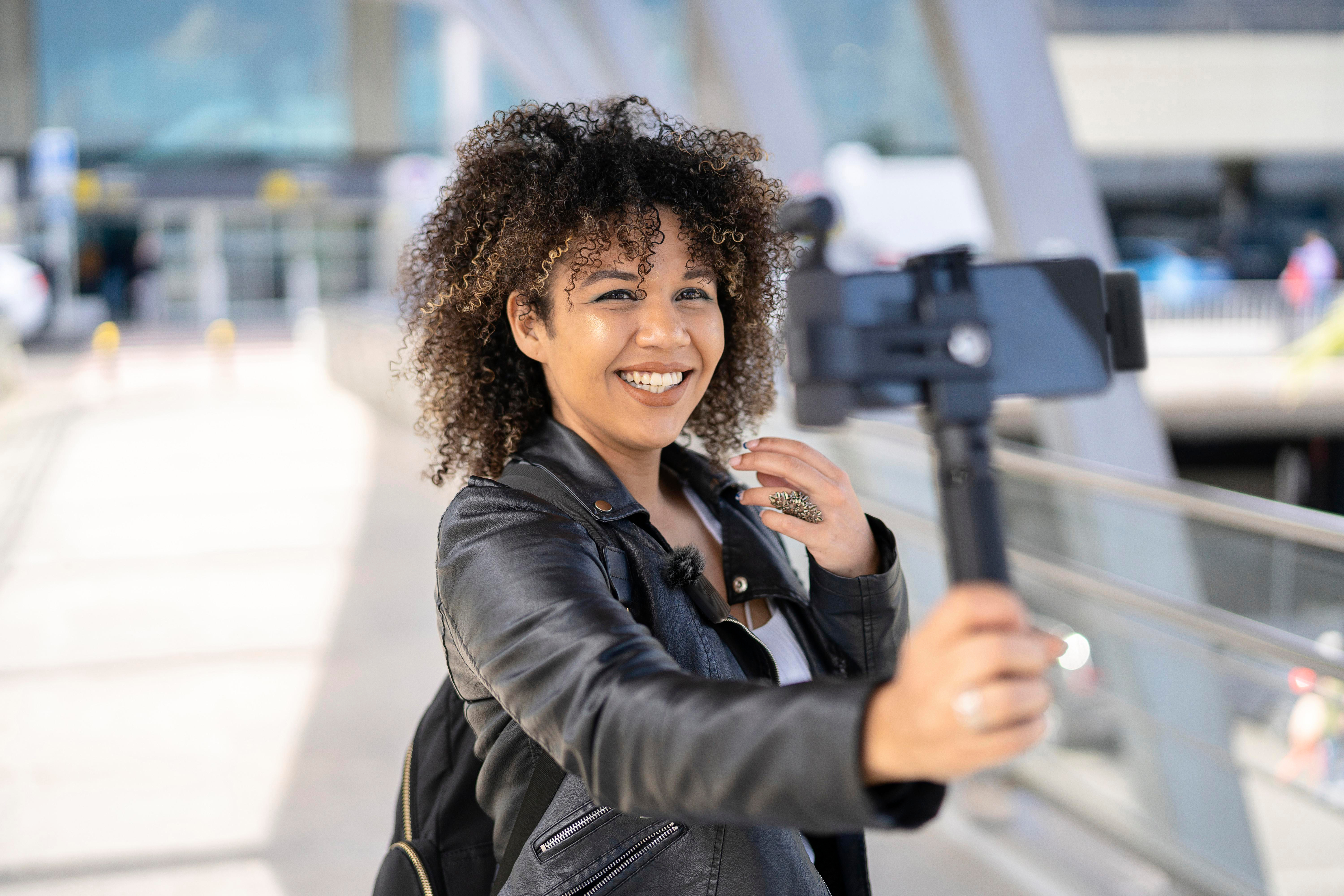 Frau in schwarzer Jacke hält Telefon auf Selfie-Stick, lächelt. Lockiges Haar und Ring sichtbar.
