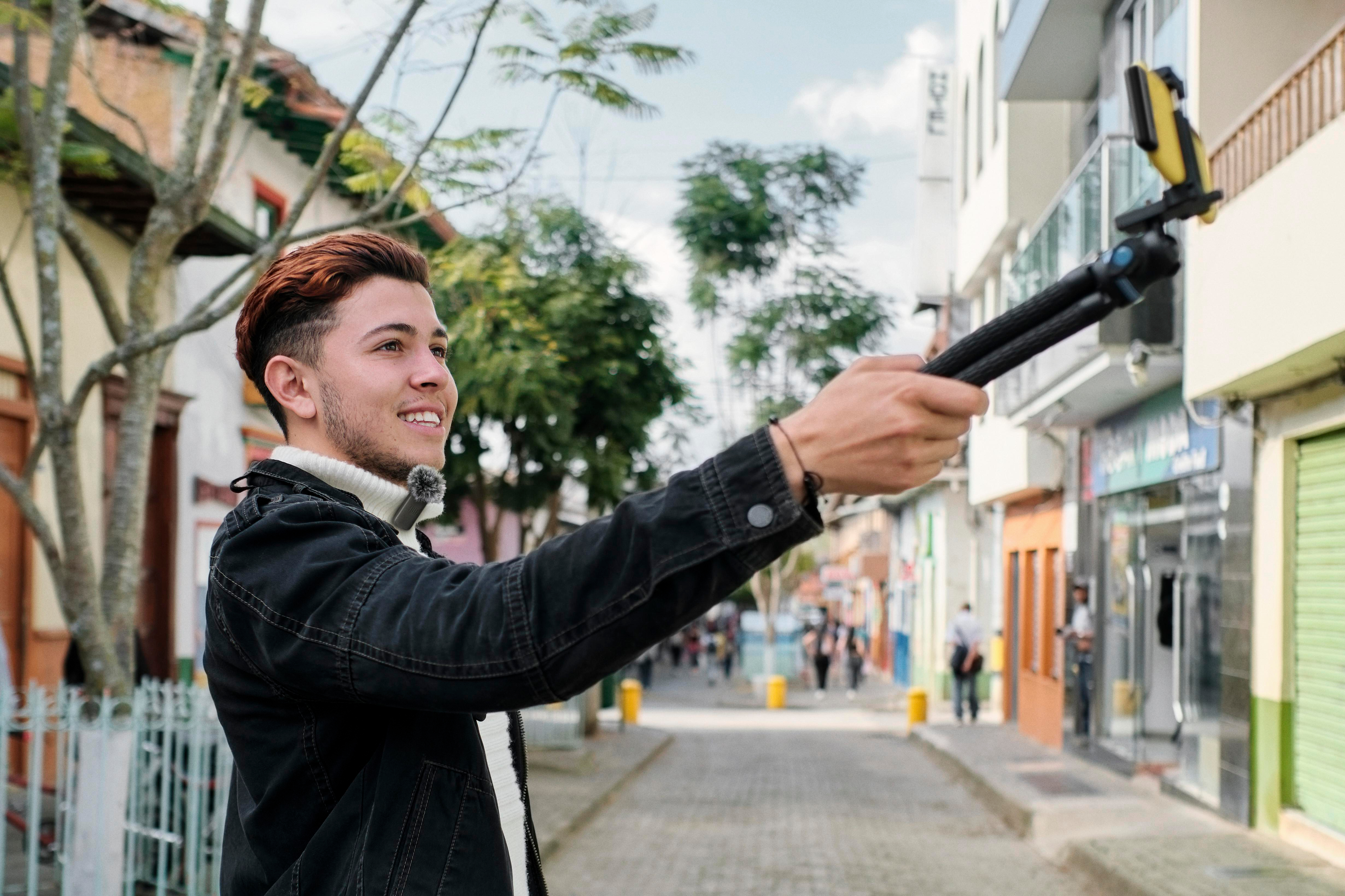 Junger Mann mit Telefon auf Stativ, lächelnd. Straße mit Geschäften und Menschen im Hintergrund.
