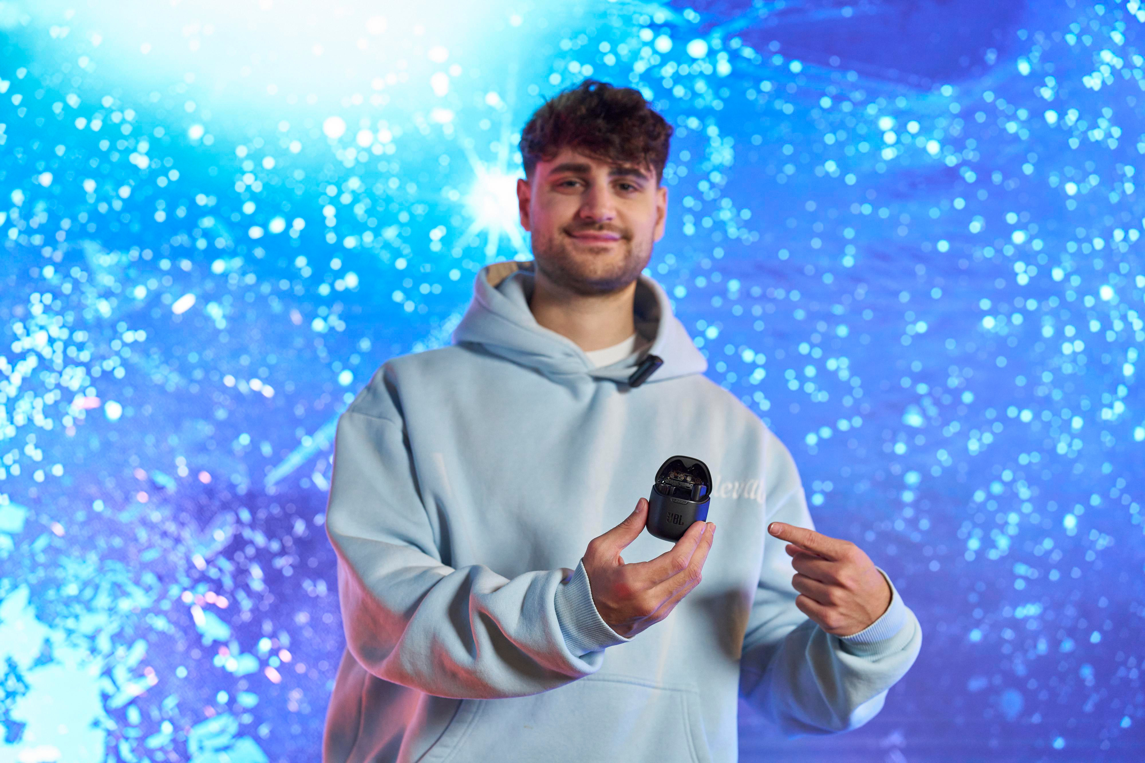 Mann mit hellblauem Hoodie hält schwarzen JBL-Kopfhörer-Etui, zeigt mit dem Finger, blauer Hintergrund.